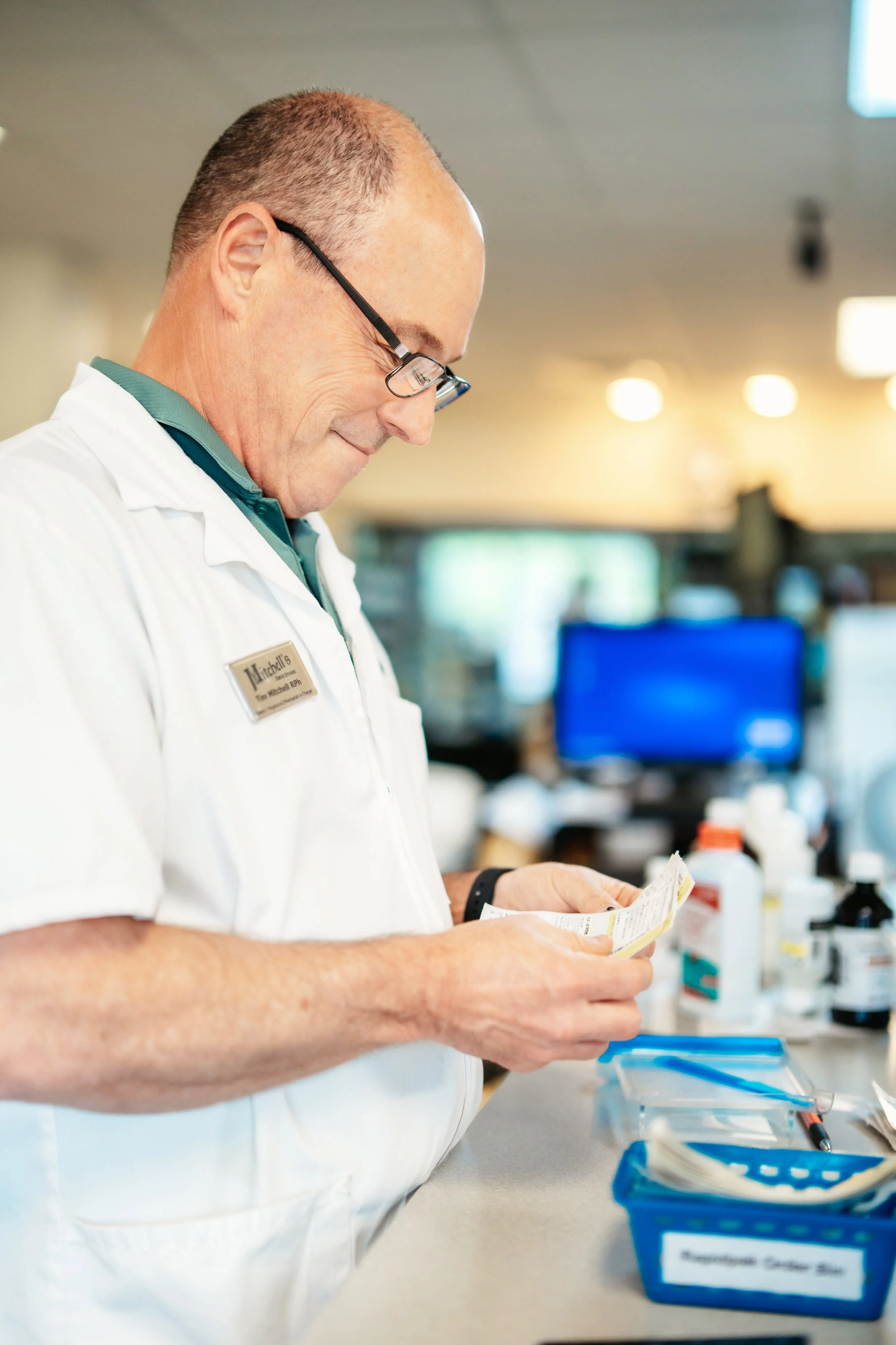 A male healthcare professional wearing glasses, a white uniform, and a name tag, is looking at a medical or prescription label, standing at a counter with medication bottles and organizing bins in the background.