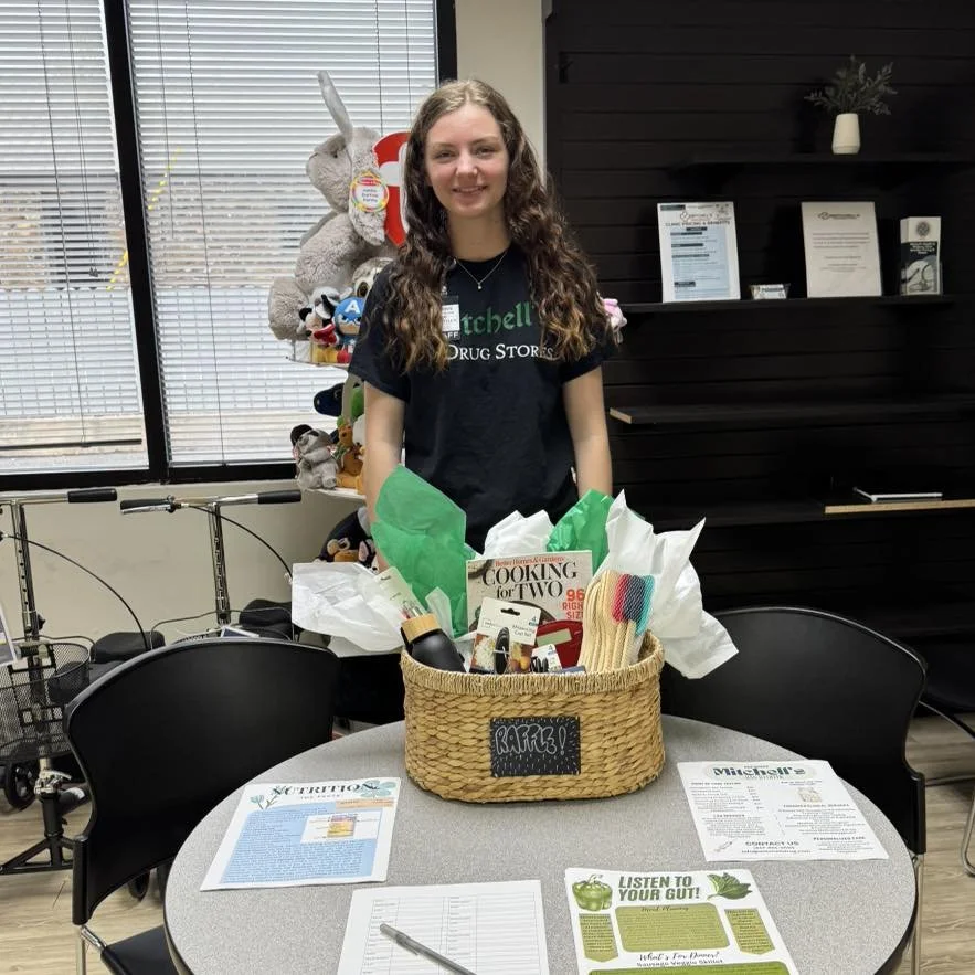 A young woman standing behind a table with a woven basket filled with various items, in a room with large windows, black chairs, and shelves with framed documents and decorative items.