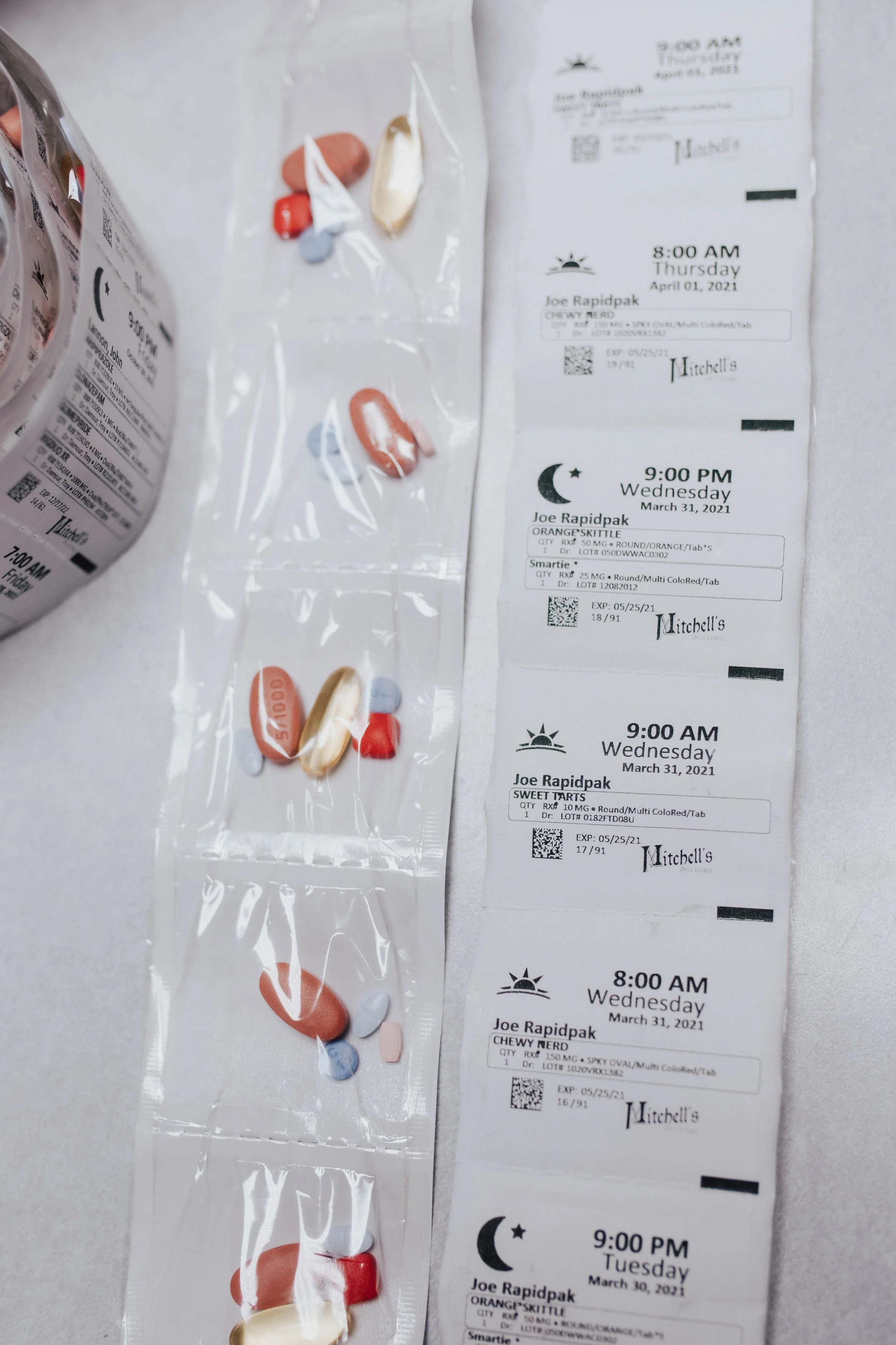 A clear plastic bag containing assorted pills, including red, blue, pink, and white capsules and tablets, resting on a surface alongside six prescription receipts for medication pickup, each with details such as time, date, patient name, medication name, and pharmacy information.
