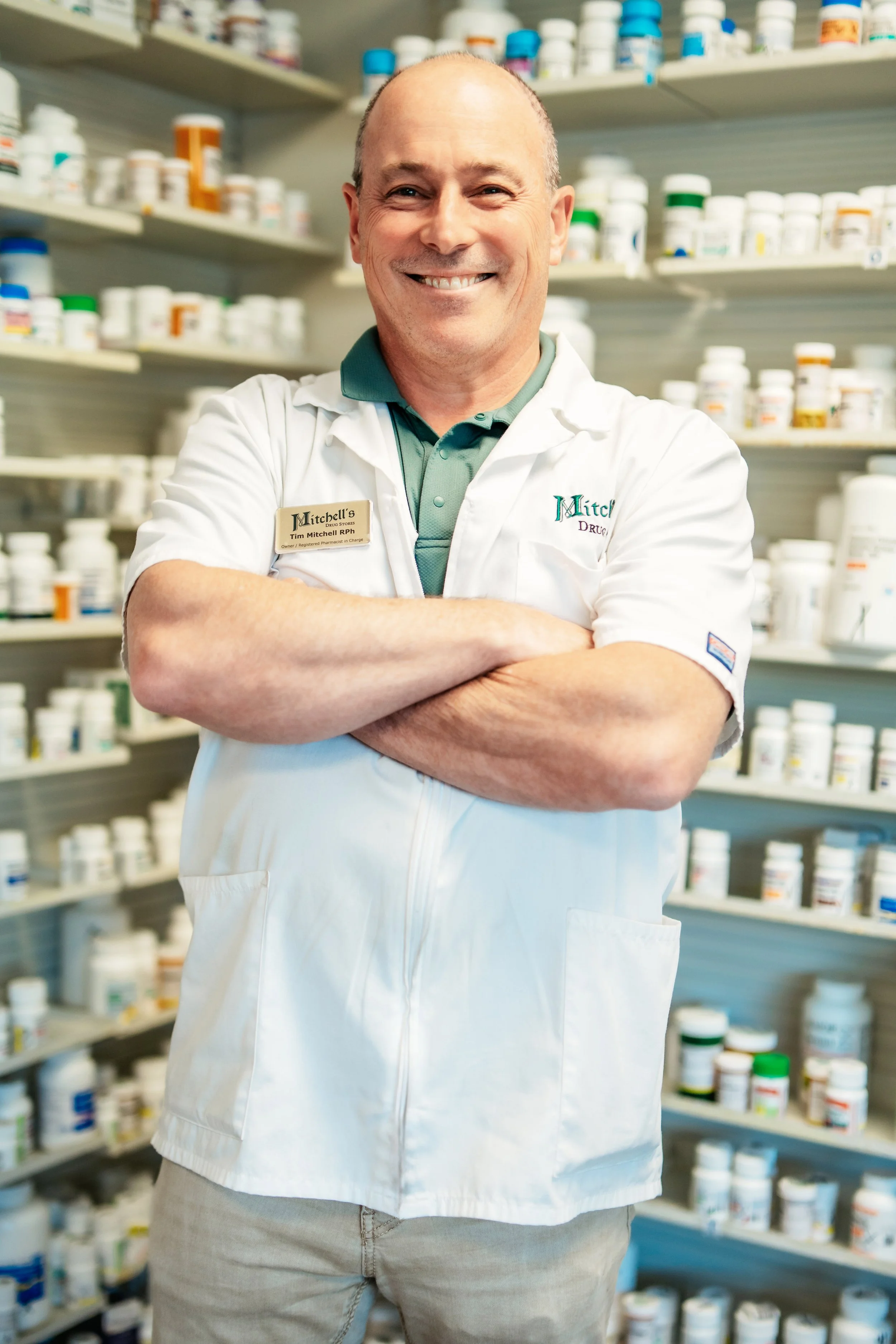 A smiling man wearing a white lab coat with a name tag, standing with arms crossed in front of shelves filled with medication bottles.