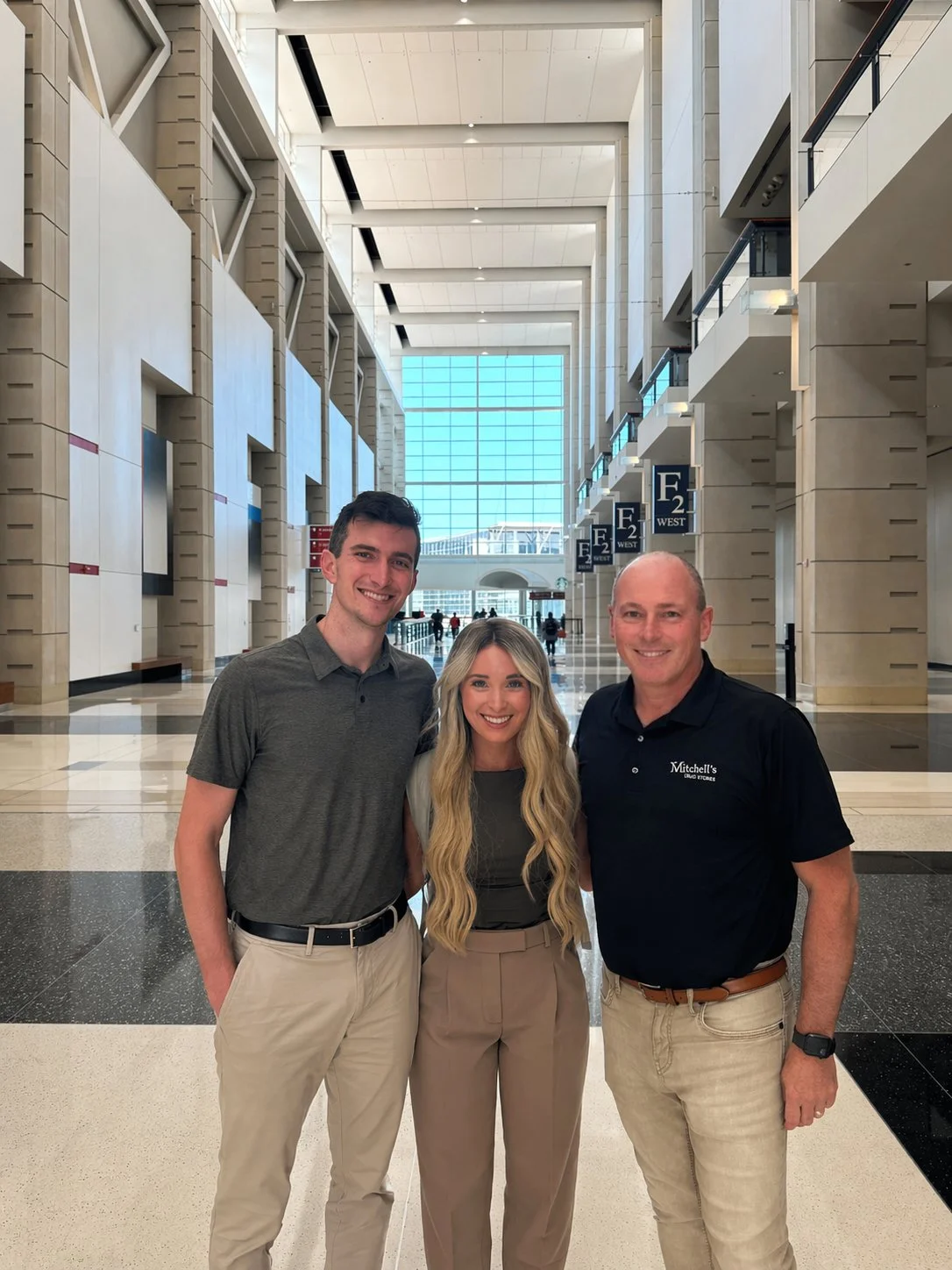 Three people standing together inside a large, modern building with high ceilings and large windows, smiling at the camera.