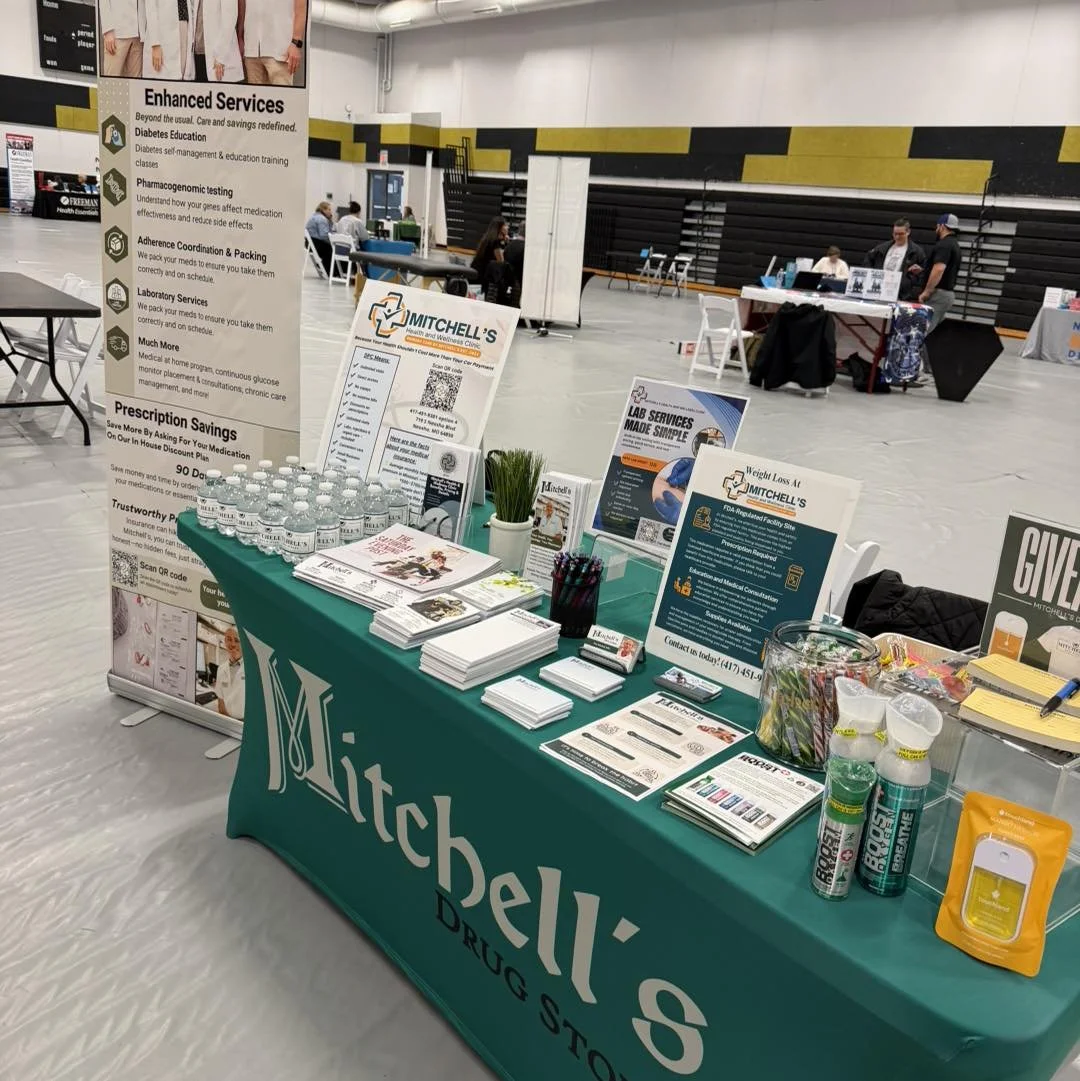 Table covered with a green tablecloth labeled 'Mitchell's Drug Store' displaying brochures, bottled water, pens, and promotional items at a health fair or community event indoors.