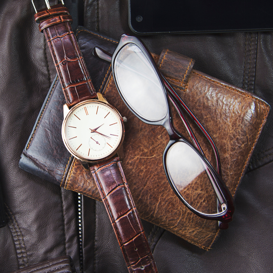 A wristwatch with a brown leather strap, a pair of eyeglasses, a brown leather wallet, and a black tablet resting on a black leather surface.