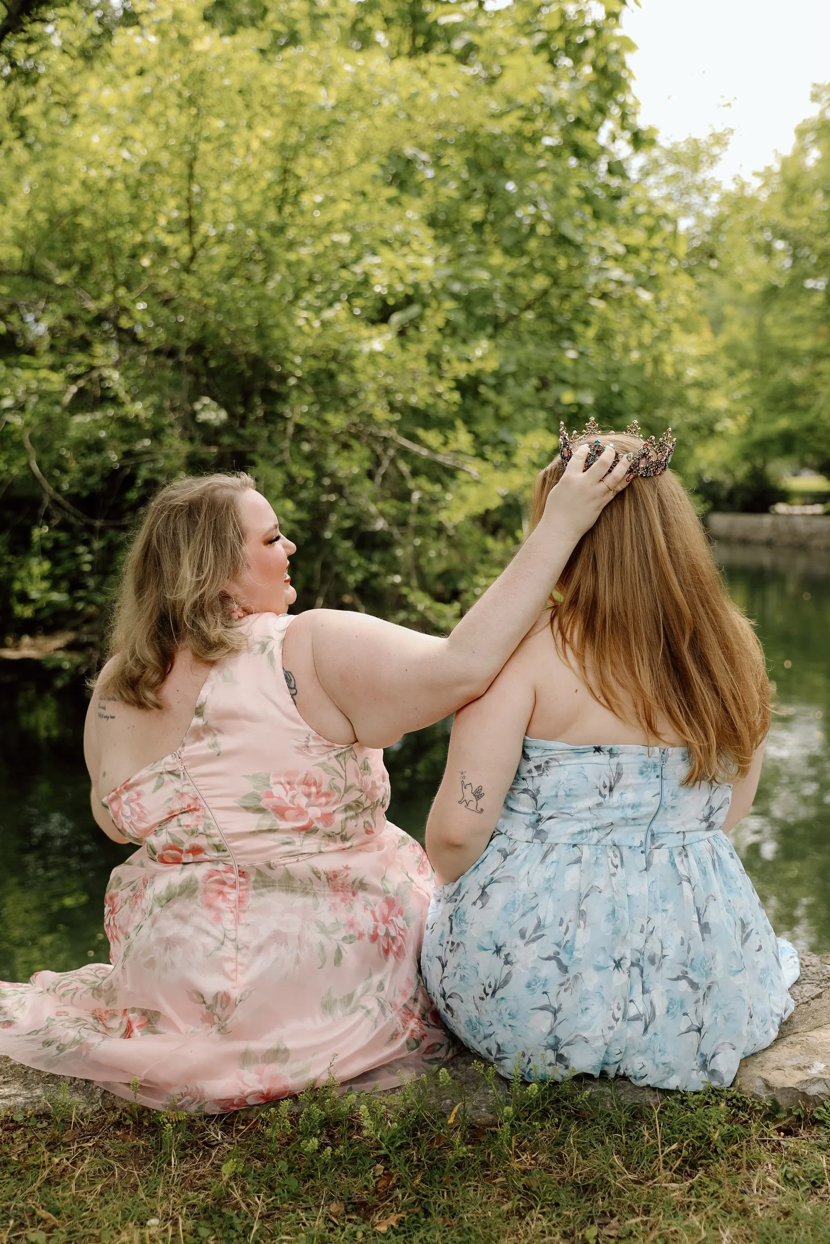 two woman wear floral dresses and look towards each other while smiling. you can see greenery in the background. the woman on the left holds up a crown to the woman on the right