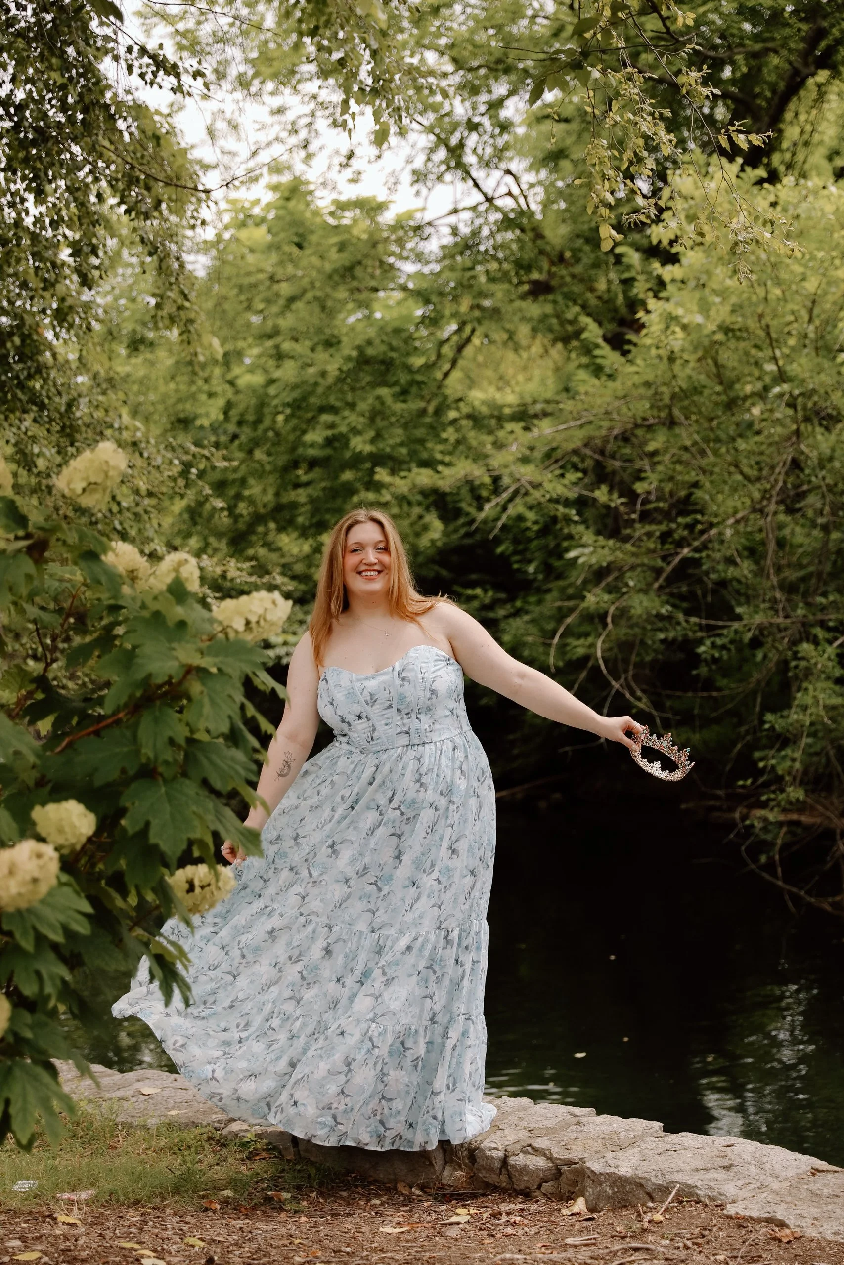 a woman stands balancing on stones near a river. you can see lots of greenery in the background. she wears a blue floral gown and holds a crown in one hand haphazardly held over the water