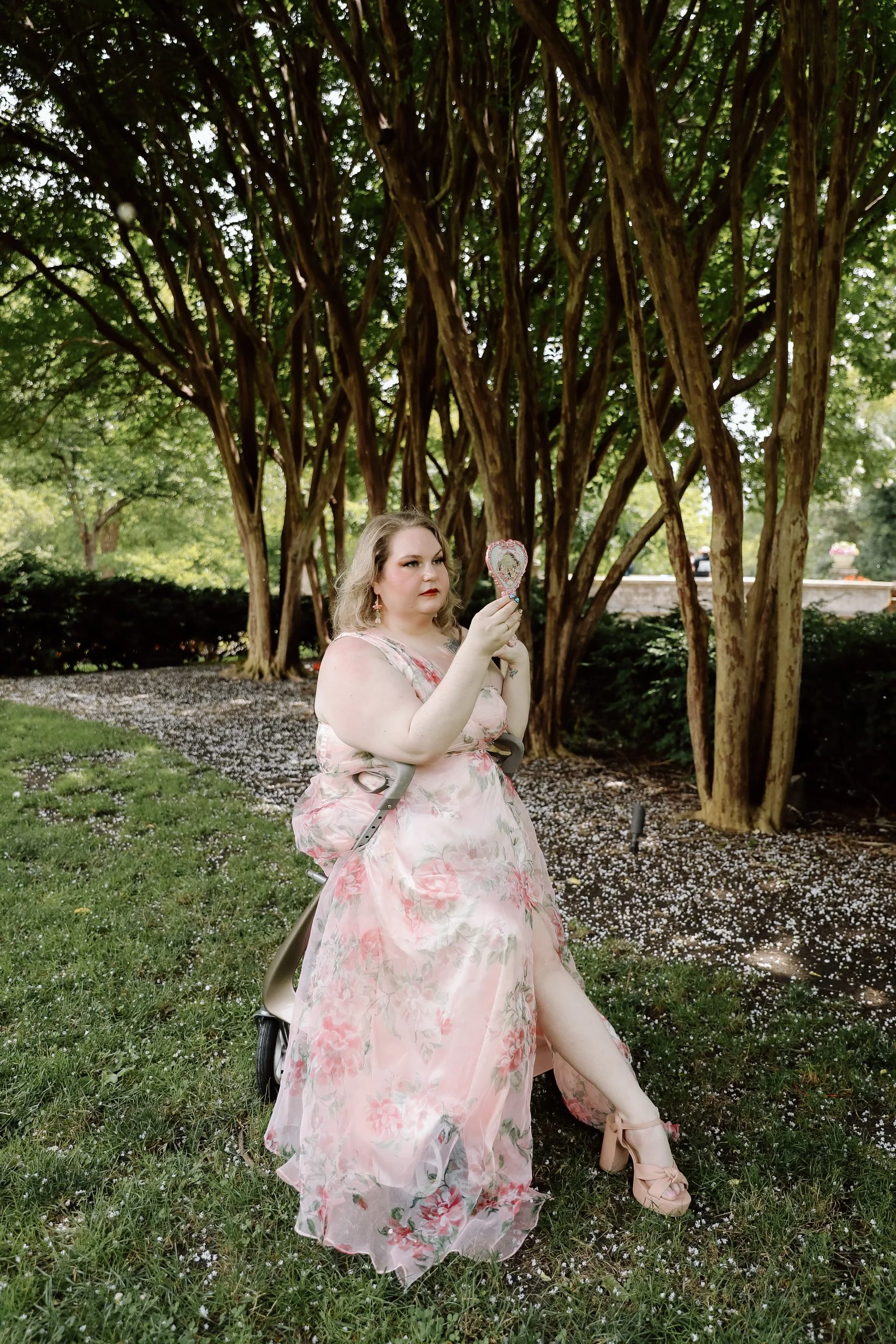 a woman sits in a rollator on grass. she wears a long floral gown and is looking into a hand held mirror