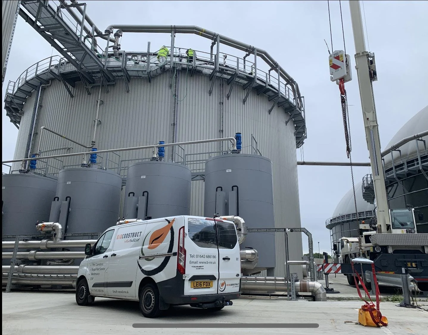 Industrial facility with storage tanks, pipes, and a crane, with a white van parked in front.