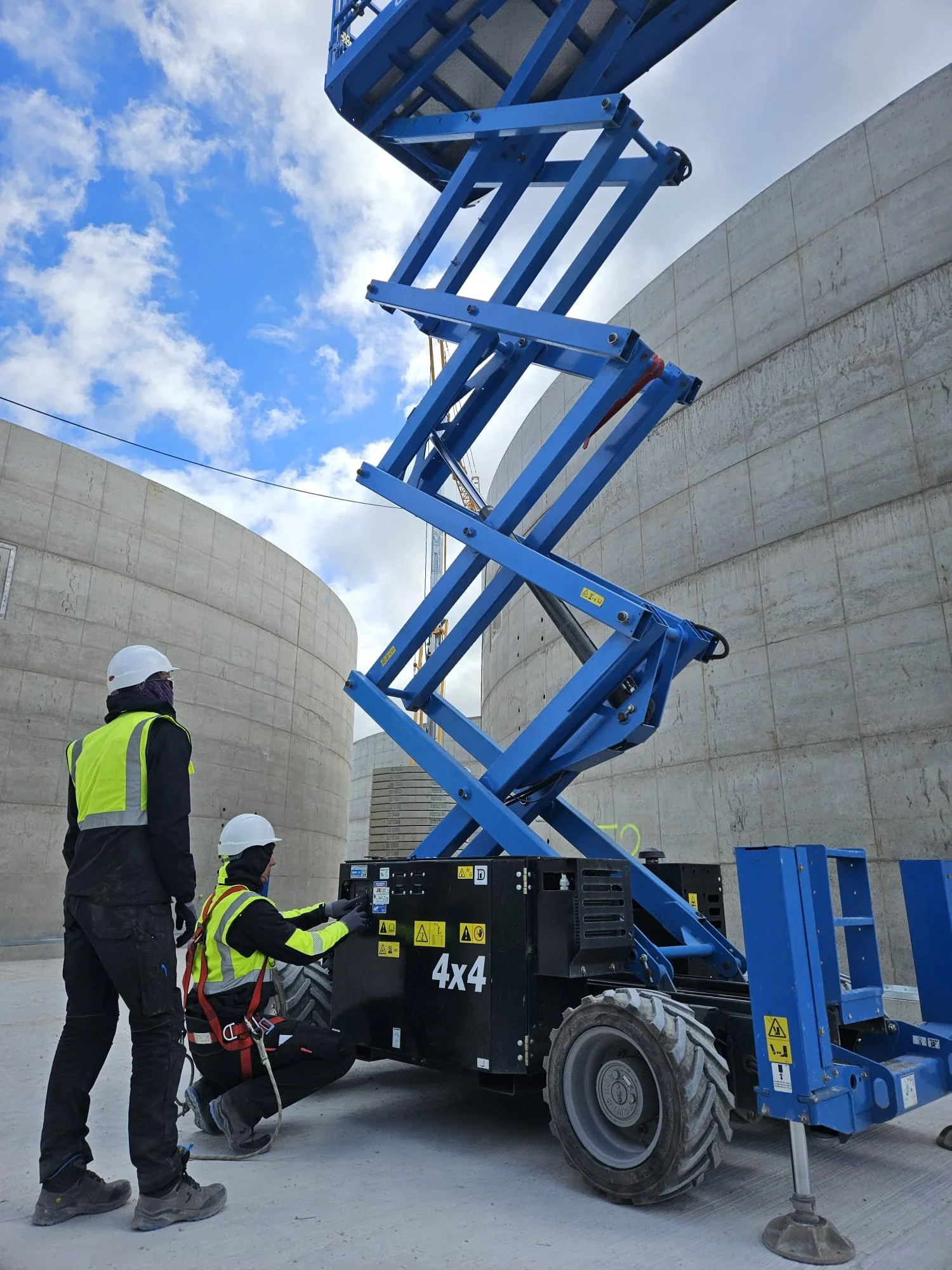 Two construction workers wearing safety helmets and vests operate a blue scissor lift in a construction site with concrete walls and a partly cloudy sky.