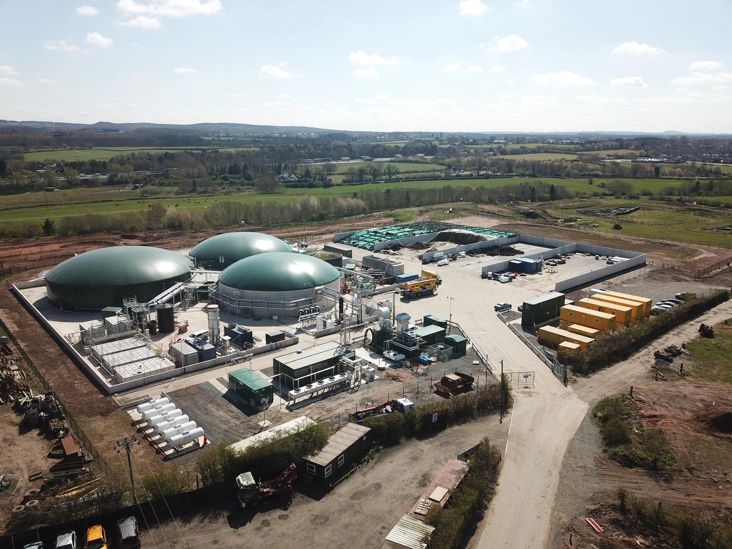 Aerial view of a biogas plant with three large green domes, surrounded by rural farmland under a partly cloudy sky.