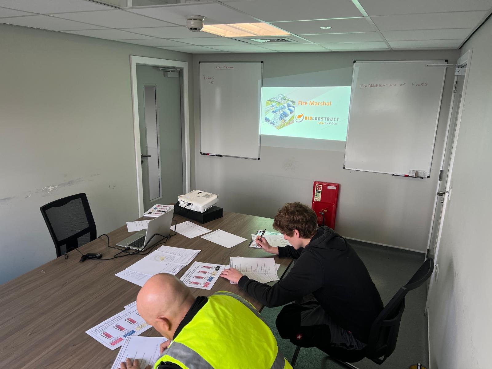 A classroom with two people reviewing documents, a whiteboard, a laptop, a projector displaying a presentation titled 'Fire Marshal' by BIOCONSTRUCT, and various papers on the table.