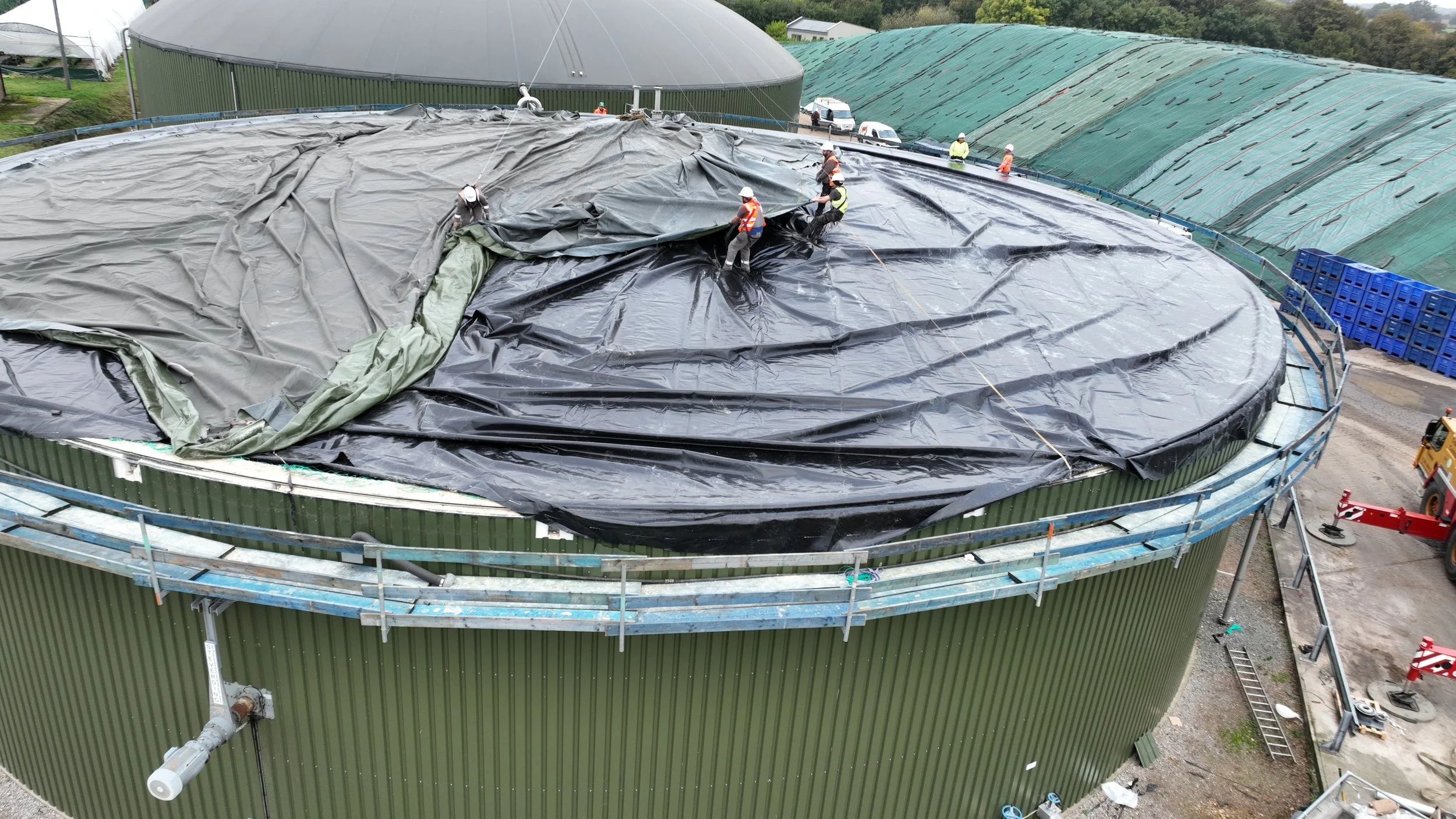 Workers installing a protective black liner or cover on the roof of a green industrial storage tank, with scaffolding around it and construction equipment nearby.