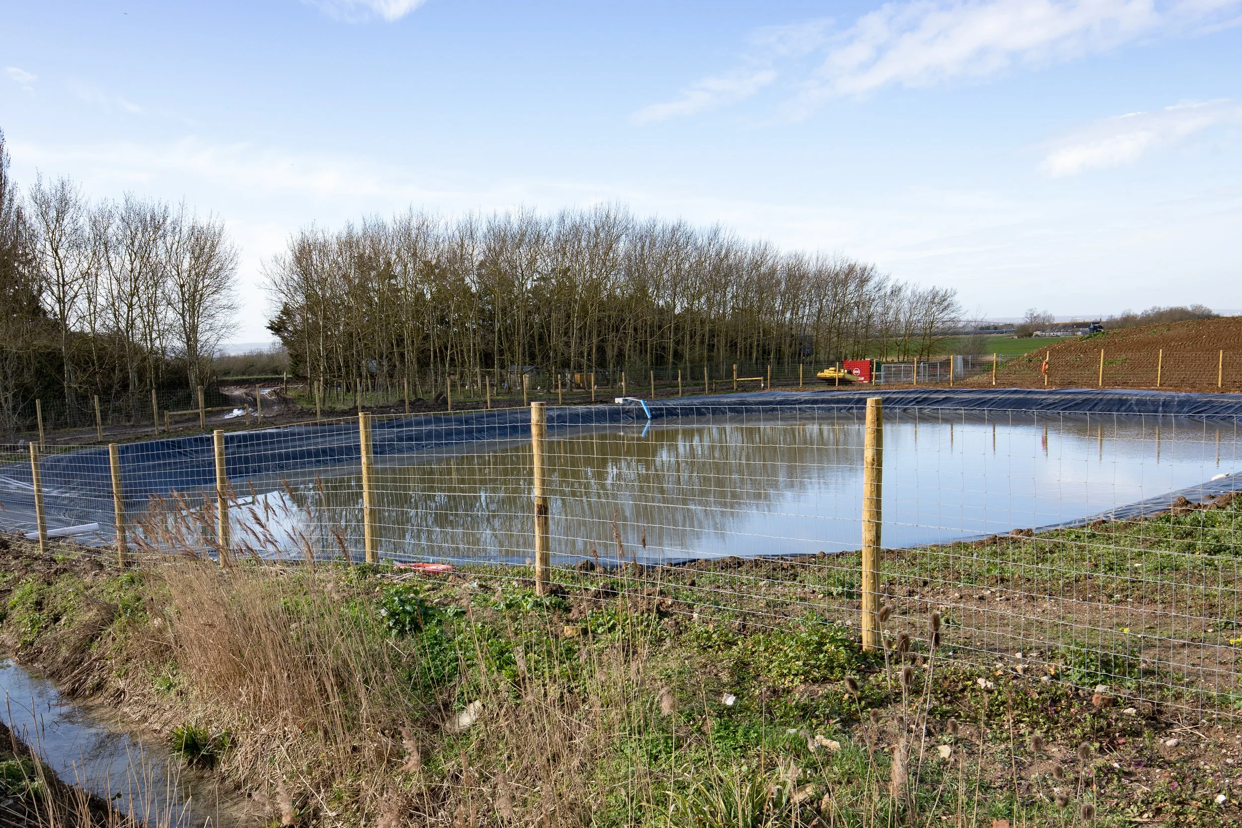 A fenced outdoor pond with black lining, surrounded by grass and trees, under a partly cloudy sky.