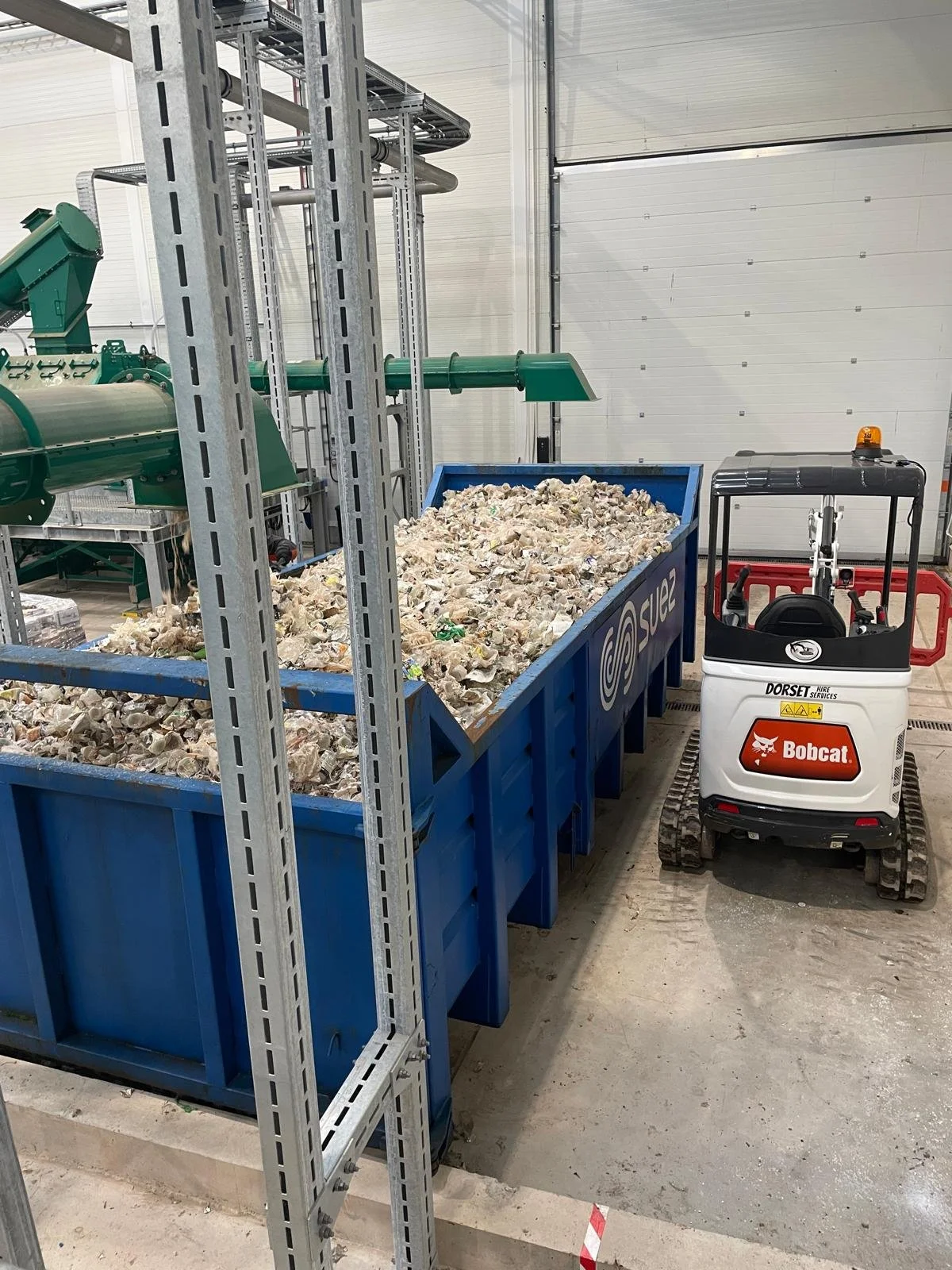 A storage container filled with crushed or shredded material, likely scrap metal or waste, next to a small Bobcat compact loader in an industrial or warehouse setting.