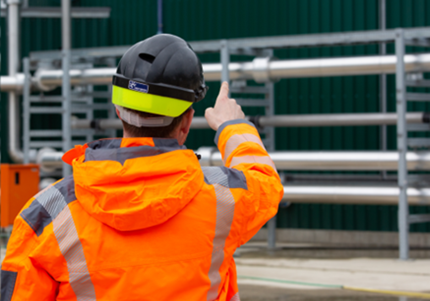 Construction worker wearing an orange high-visibility jacket and safety helmet, pointing at metal pipes on a construction site.