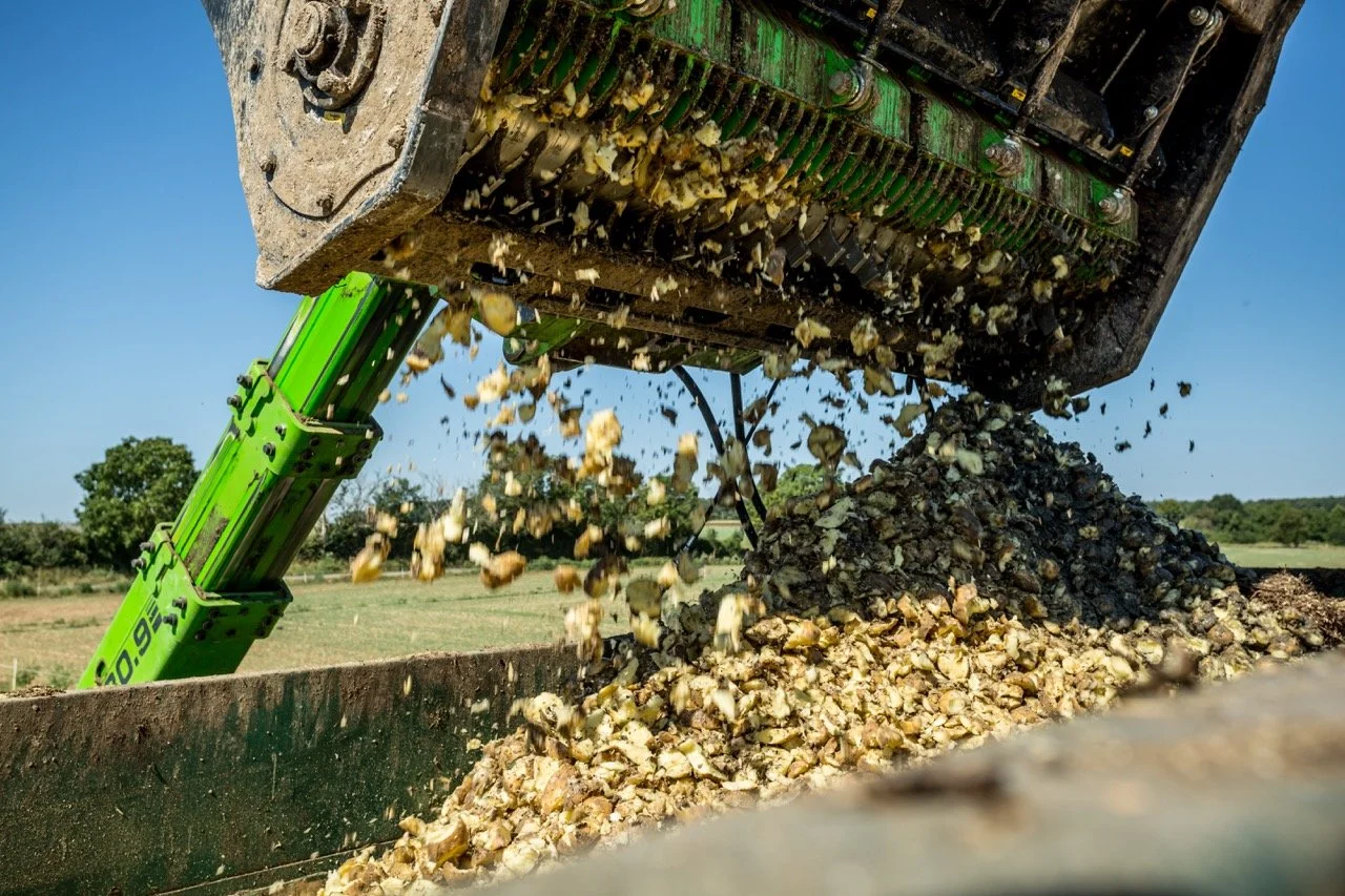 Close-up of a green agricultural harvester ejecting a mixture of crushed stone or gravel onto a pile in an open field.
