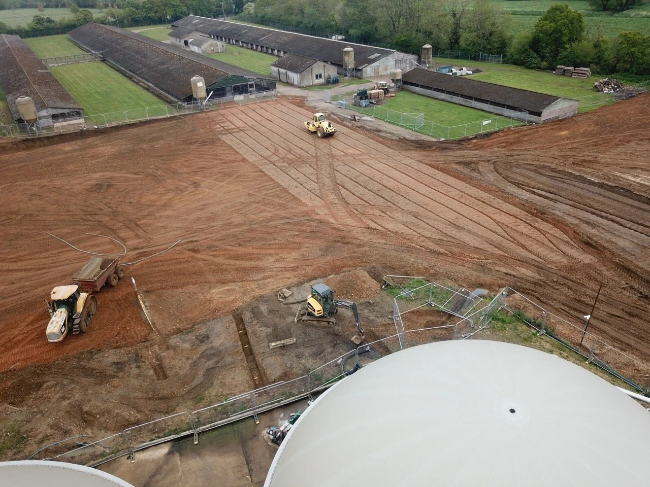 Construction site with bulldozers and a roller compactor preparing land, adjacent to farm buildings and green fields.