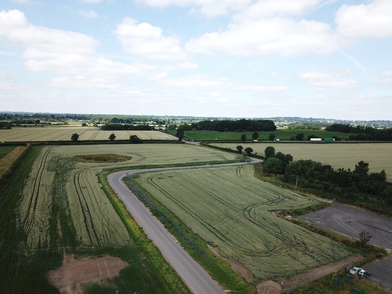 Aerial view of farmland with green fields, a winding road, and scattered trees under a partly cloudy sky.