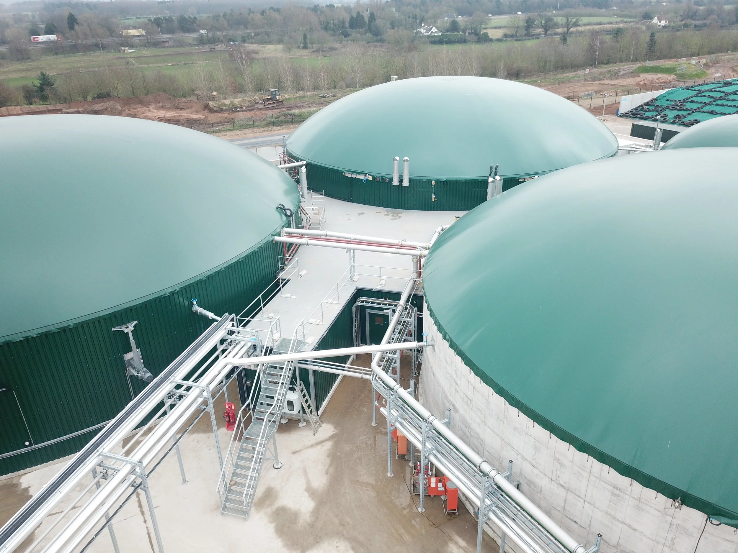 Industrial storage tanks with green domed roofs connected by metal pipes and stairs, set in a rural area with fields and trees in the background.