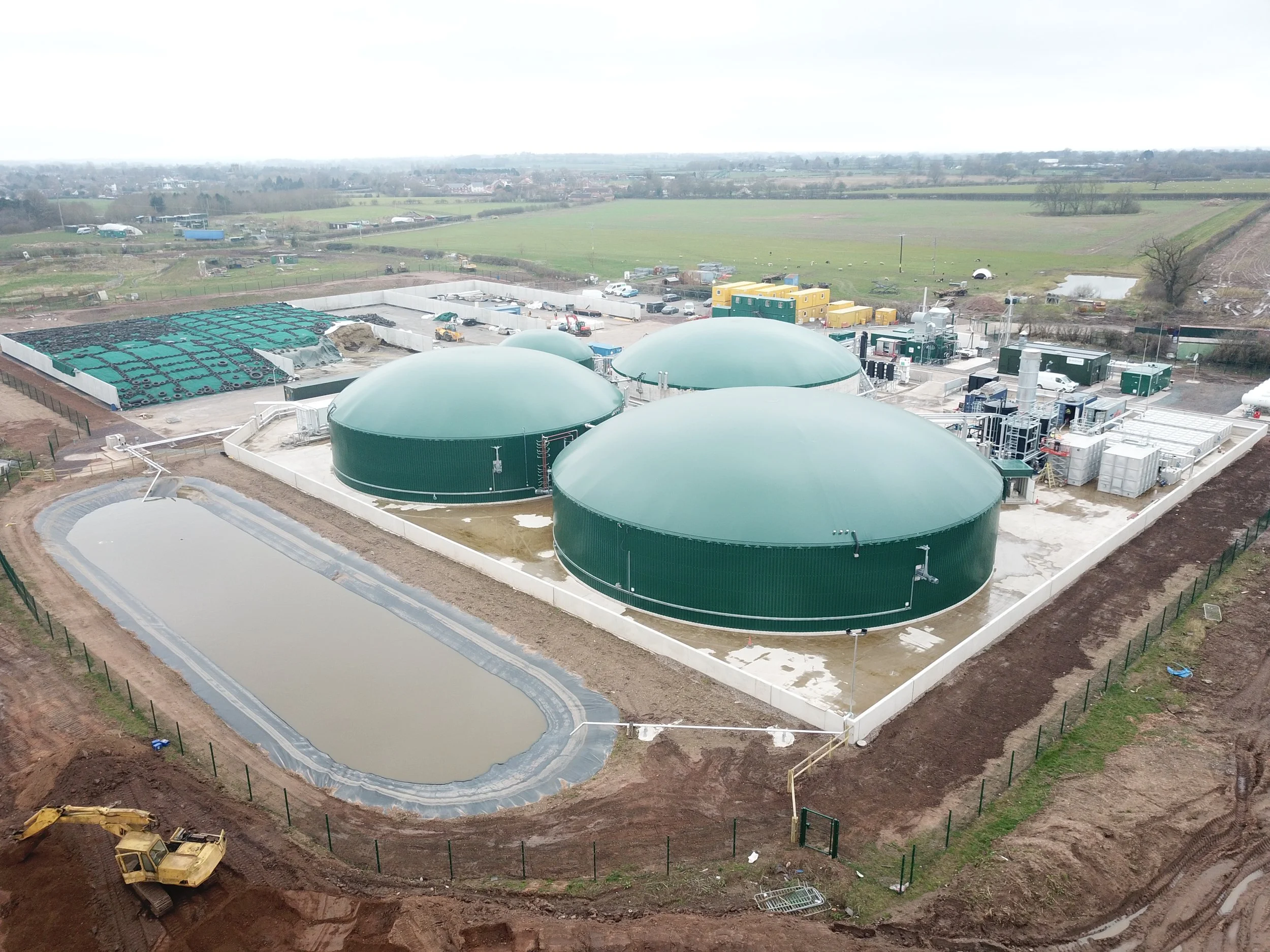 Large green biogas or methane digesters at an industrial facility on a farm with a pond and farmland in the background.
