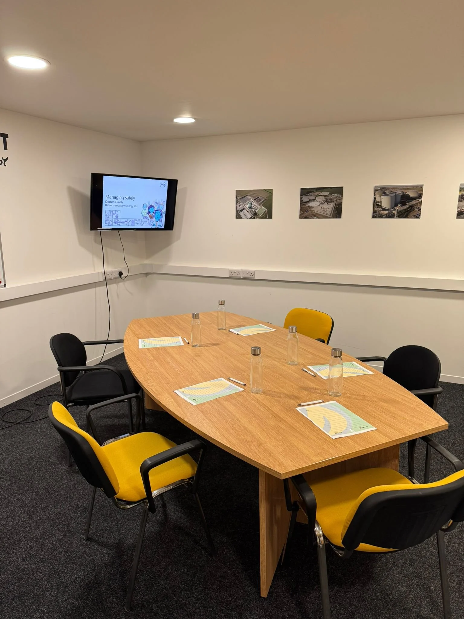 A conference room with six yellow and black chairs around a wooden oval table, each with a glass water bottle and a folder. A wall-mounted screen displays a presentation titled "Managing safety" and several framed aerial photos hang on the white walls.