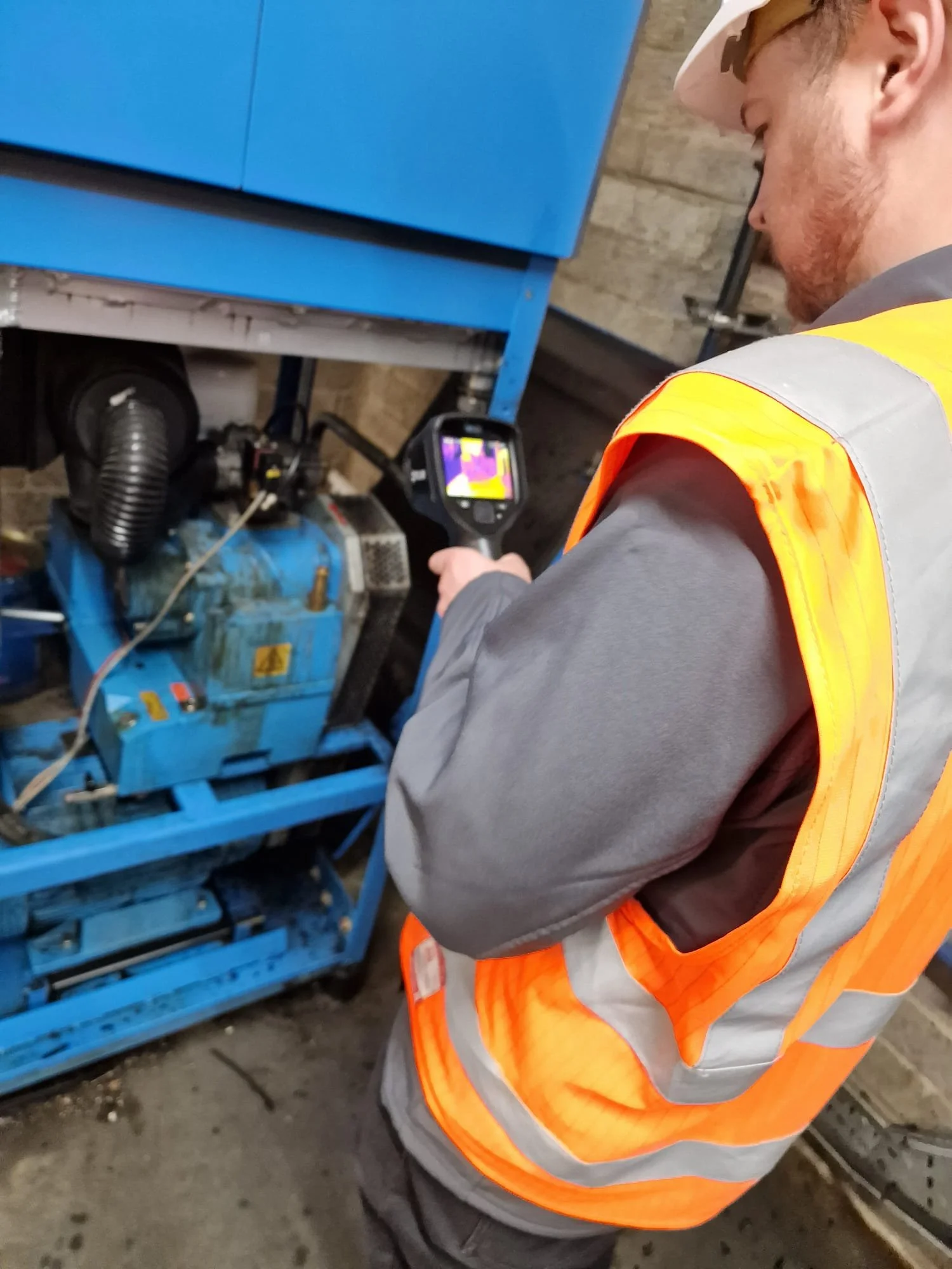 A technician in a gray shirt and orange safety vest uses a thermal imaging camera to inspect a blue industrial machine.