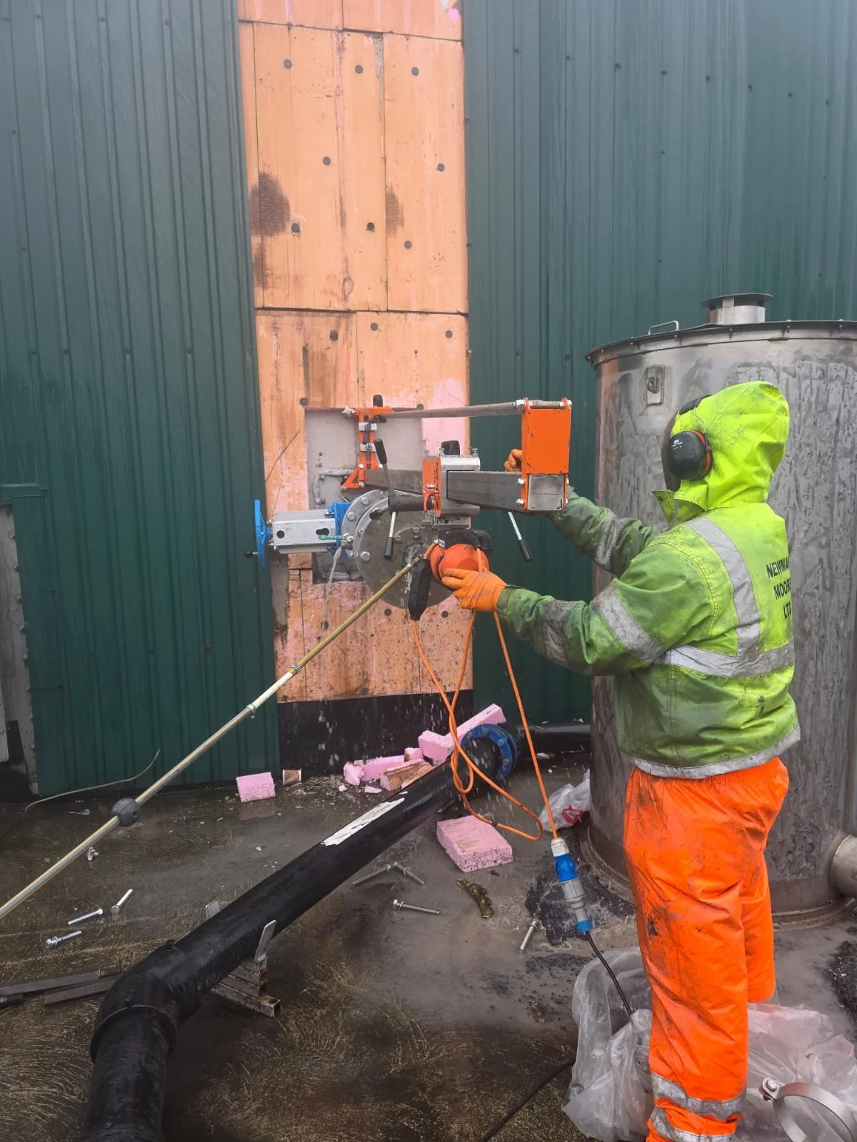 Worker in high-visibility safety gear operating a core drilling machine on the side of a building with pink insulation foam, green and orange walls, and a large metal tank nearby.