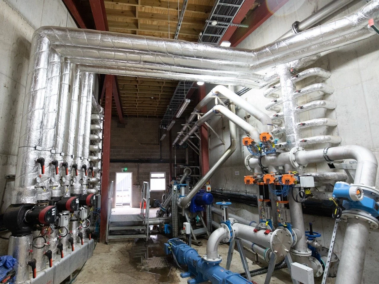 Industrial mechanical room with numerous insulated pipes, valves, and fittings for heating or cooling systems, with an open doorway in the background.