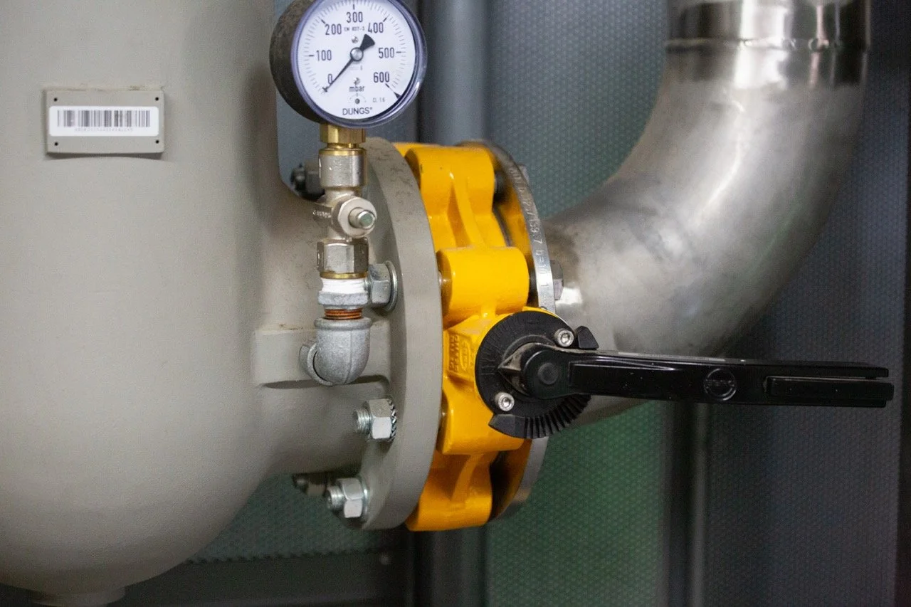A close-up view of industrial equipment, including a pressure gauge, metallic pipes, a yellow valve, and a black adjustment lever.