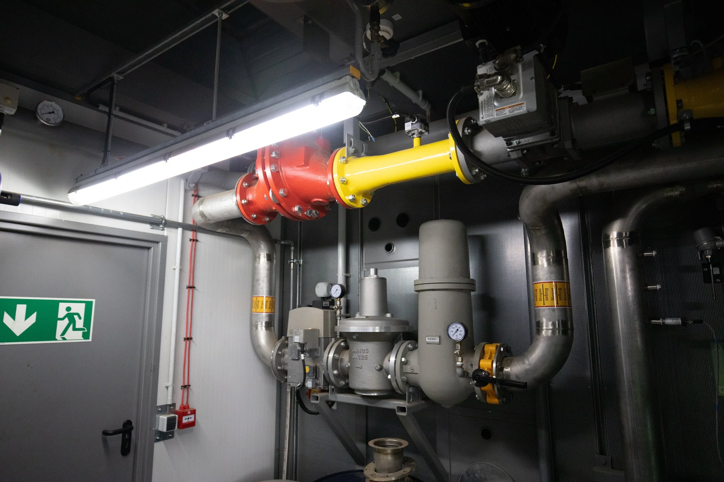 Industrial pipes and valves with gauges, including a red and yellow pipe, in a mechanical room.