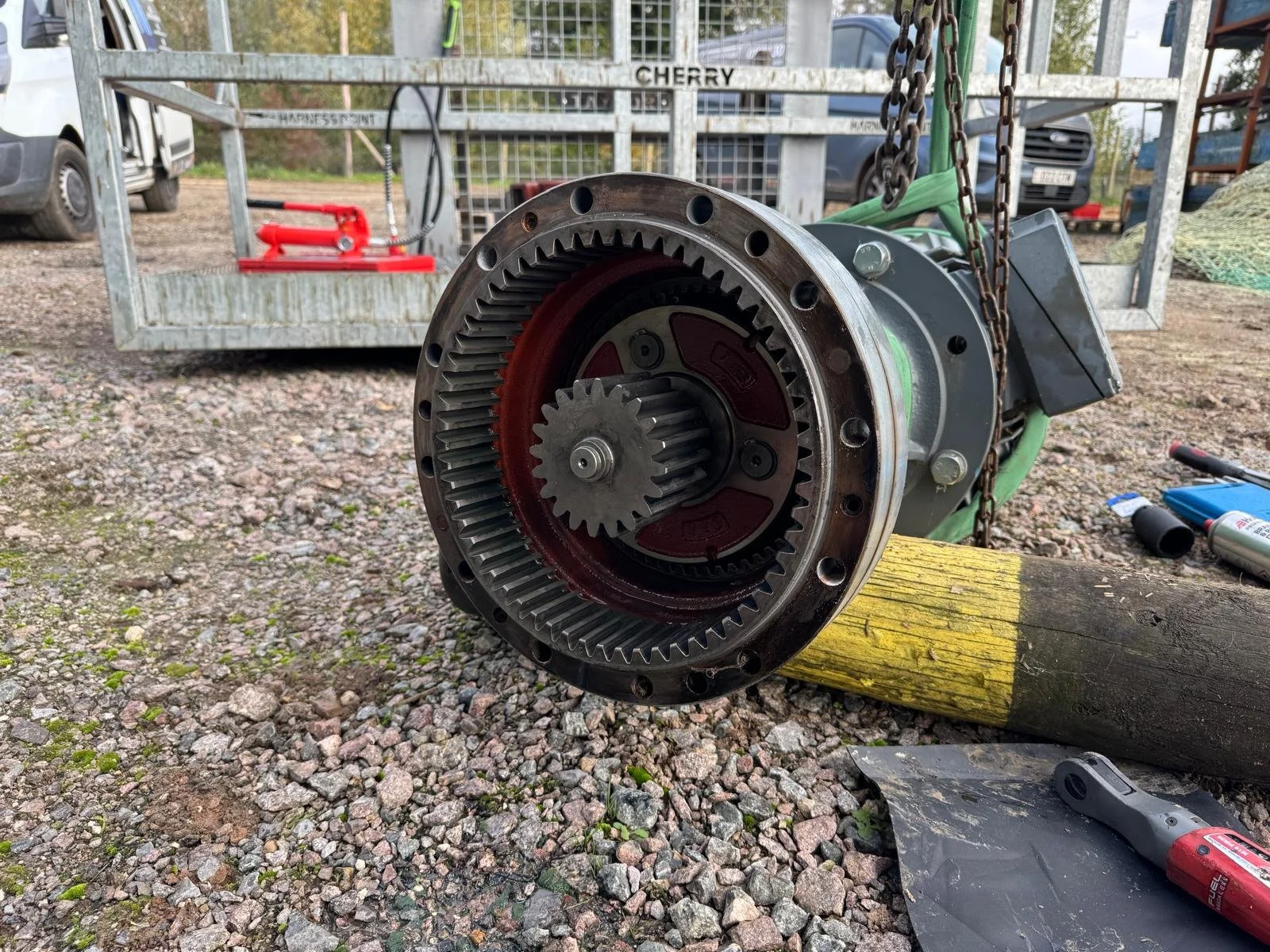 Close-up of a large mechanical gear assembly resting on a rough gravel surface outdoors, with tools and equipment in the background.
