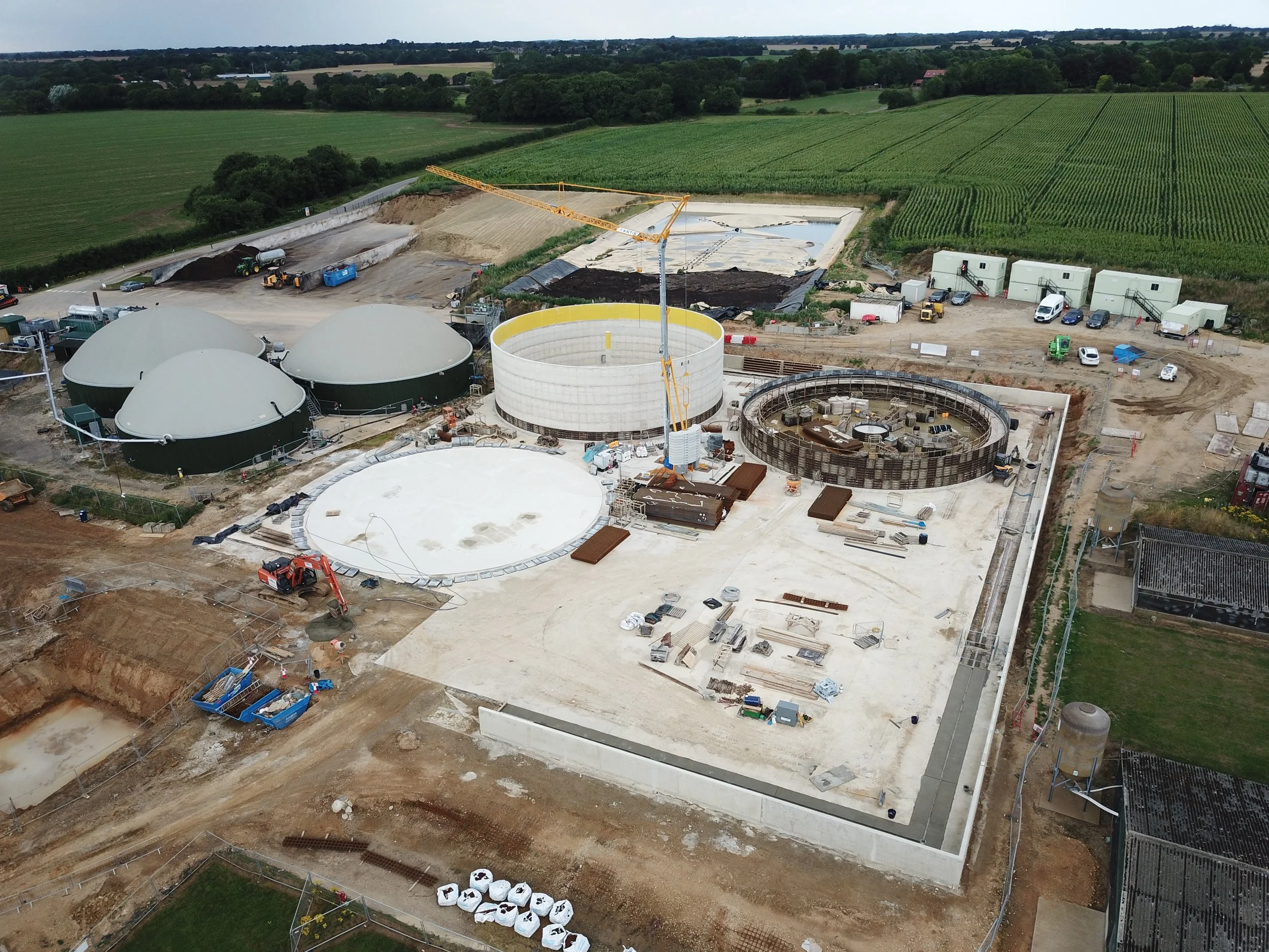 Aerial view of a construction site for a water treatment plant with large storage tanks, a tower crane, and surrounding farmland.