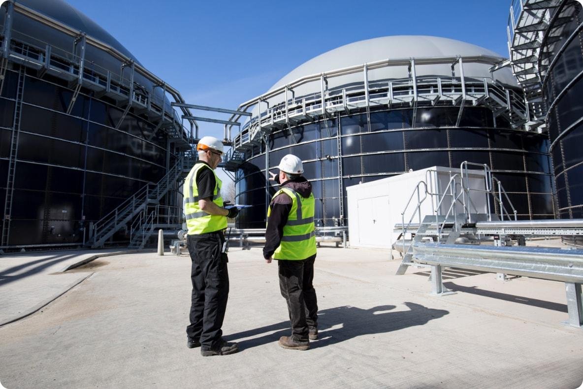 Two workers wearing safety vests and helmets standing outside a large industrial facility with black tanks and metal walkways.