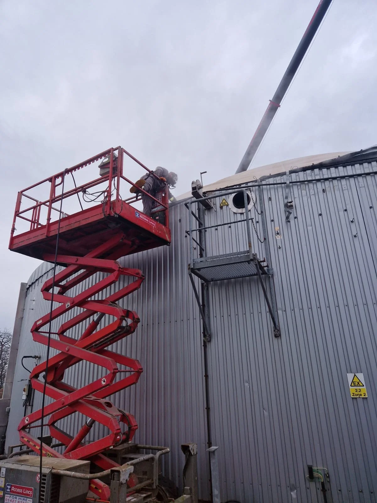 A worker on a red scissor lift inspecting or repairing an industrial building's exterior wall under a cloudy sky.