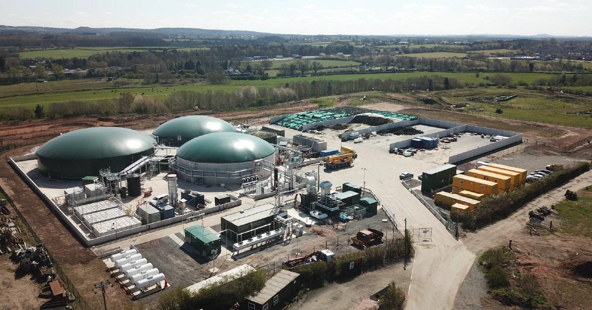 Aerial view of a biogas plant with three large green dome digesters, surrounded by storage tanks, equipment, and containers on a gravel lot, with green fields and farmland in the background.