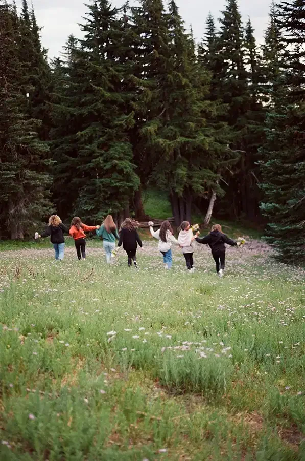Group of seven children holding hands while walking through a grassy field with wildflowers, surrounded by tall evergreen trees.