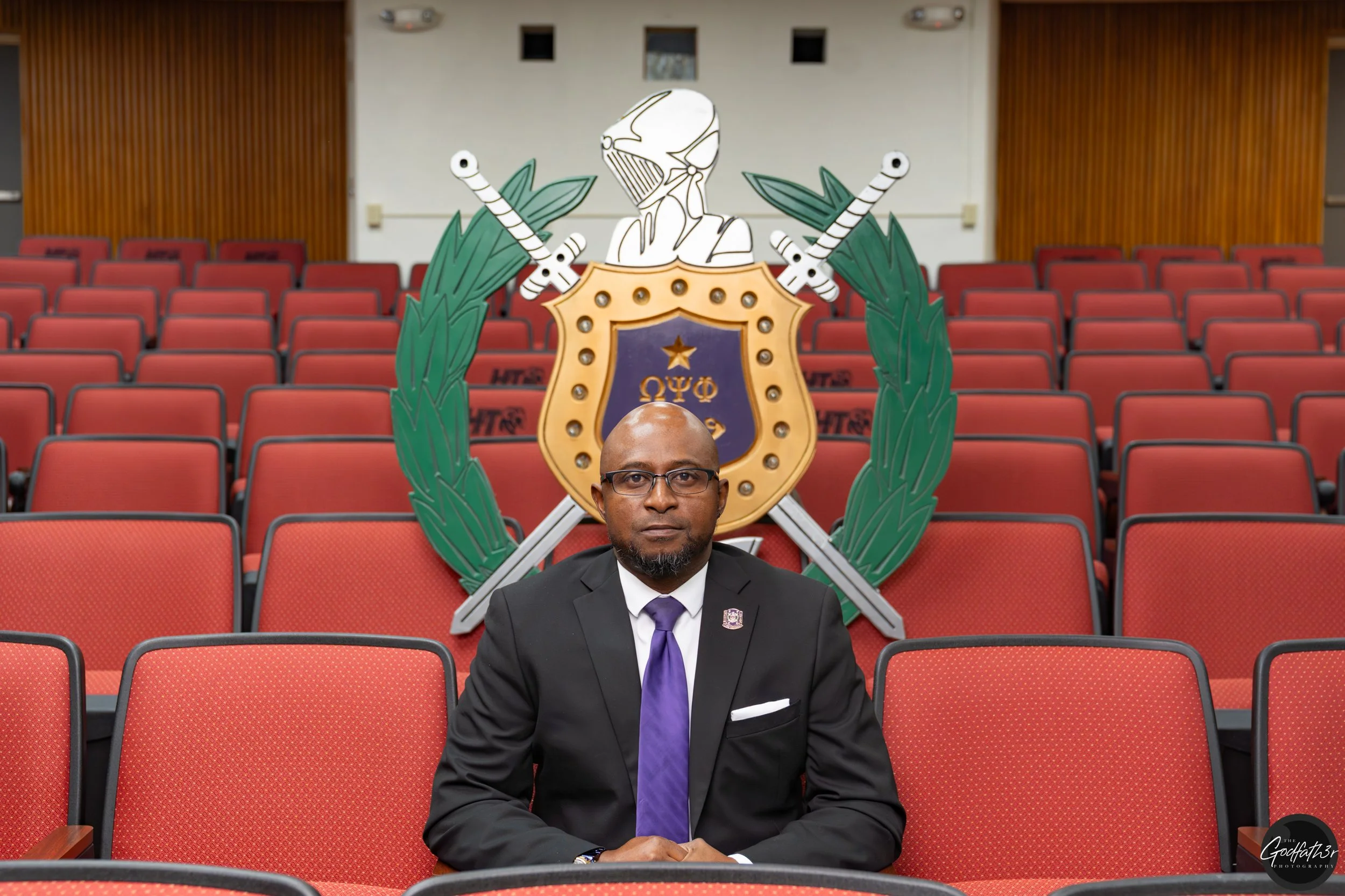 A man in a navy suit, white shirt, and blue dotted tie, with a beard and close-receding hair, posing against a neutral backdrop.