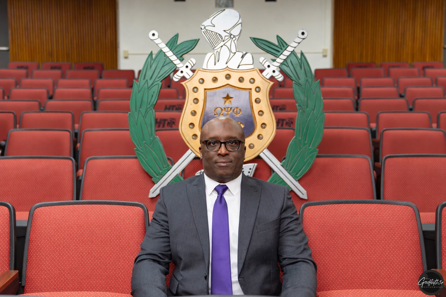 A man dressed in a black suit, white shirt, and purple tie, wearing glasses, standing against a plain light-colored background.
