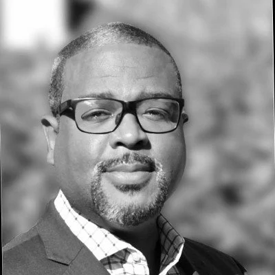 Black and white portrait of a man wearing glasses, a checkered shirt, and a suit jacket.