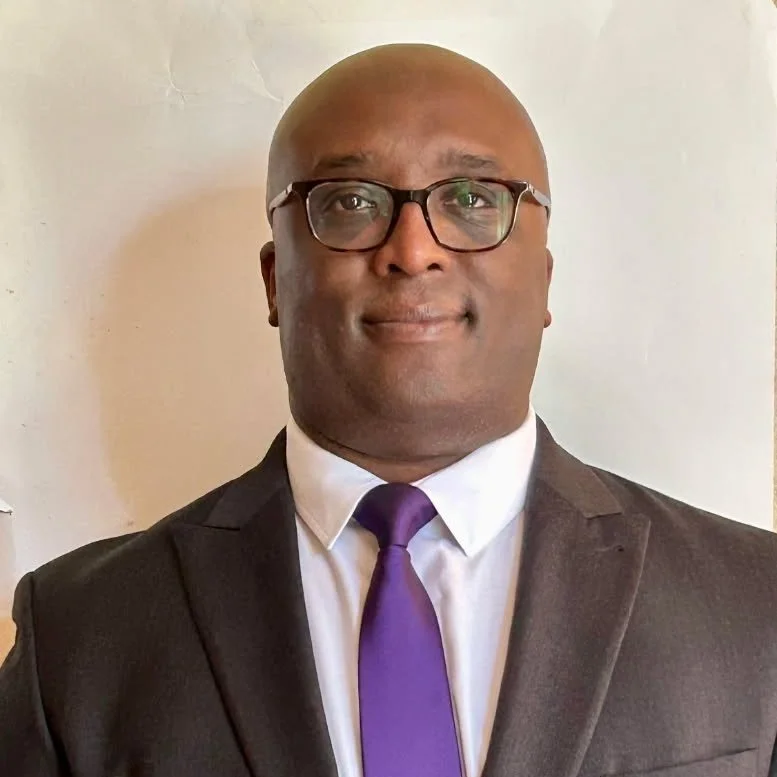 A man dressed in a black suit, white shirt, and purple tie, wearing glasses, standing against a plain light-colored background.