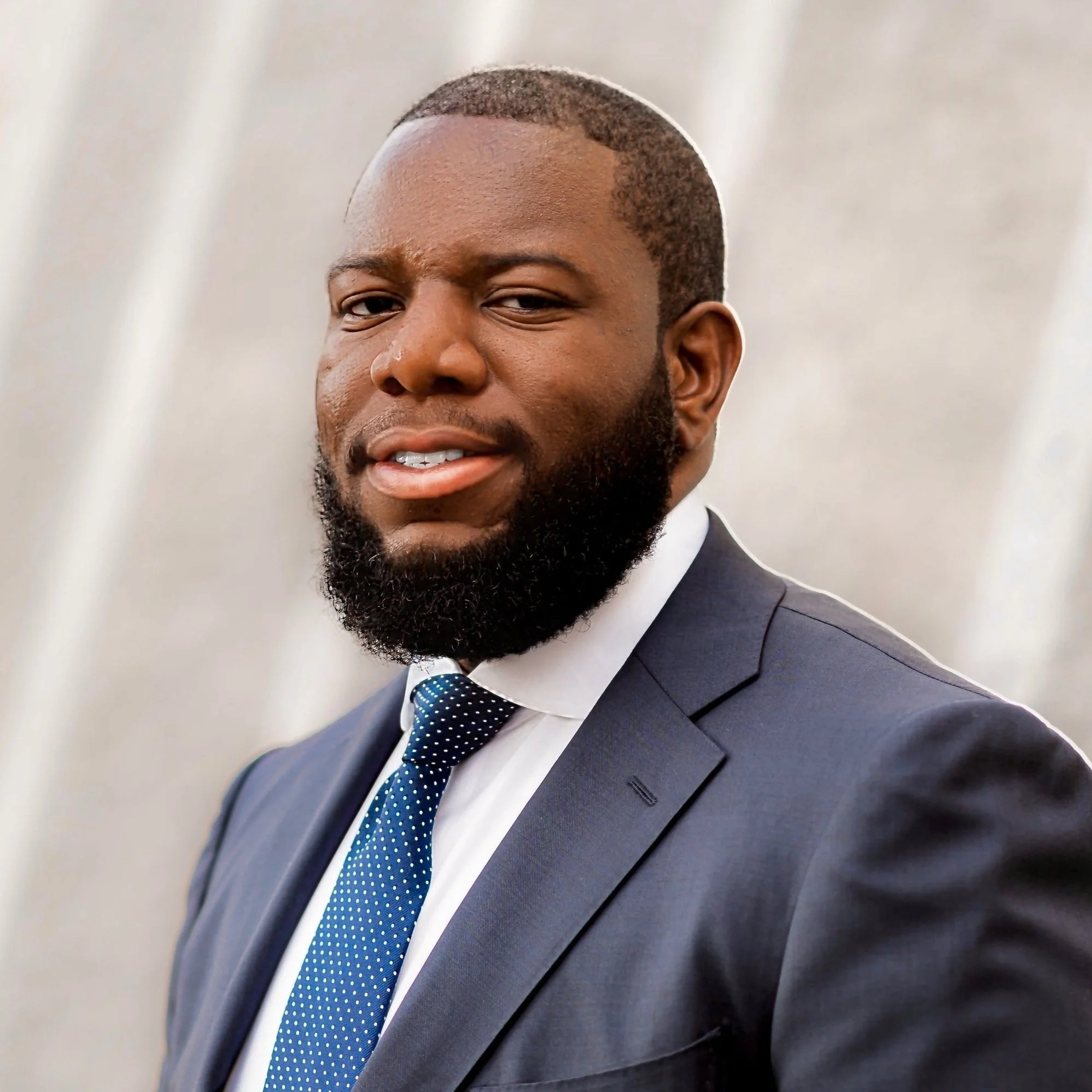 A man in a navy suit, white shirt, and blue dotted tie, with a beard and close-receding hair, posing against a neutral backdrop.