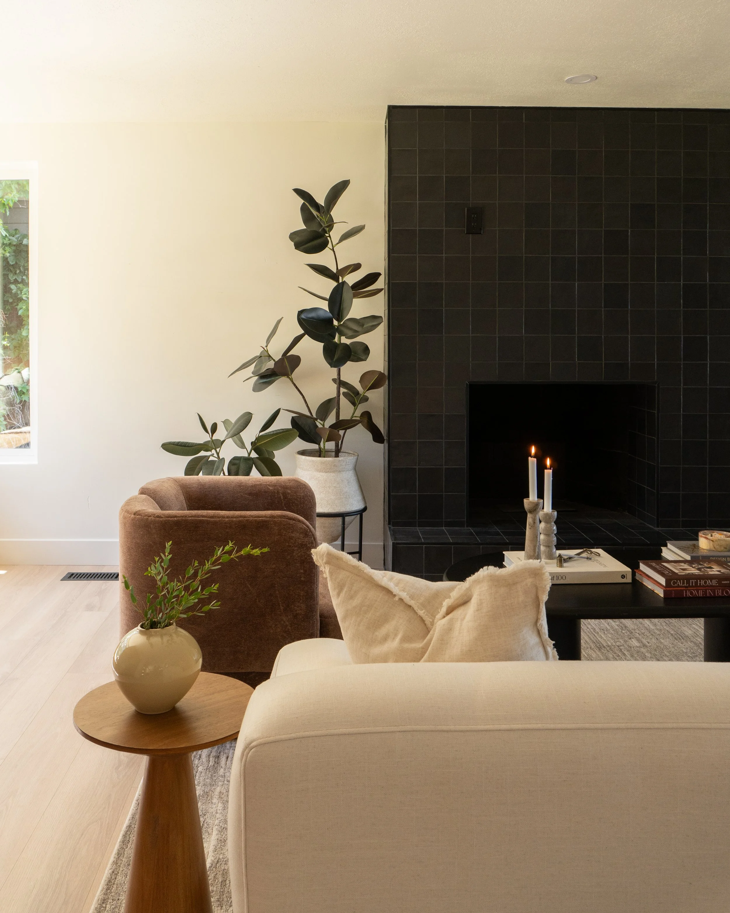 Living room with a round wooden table holding a beige vase with green plants, a beige armchair, a burgundy armchair, a black fireplace with candles, a large green plant in a white pot, a black side table with books, and a black tiled fireplace with c
