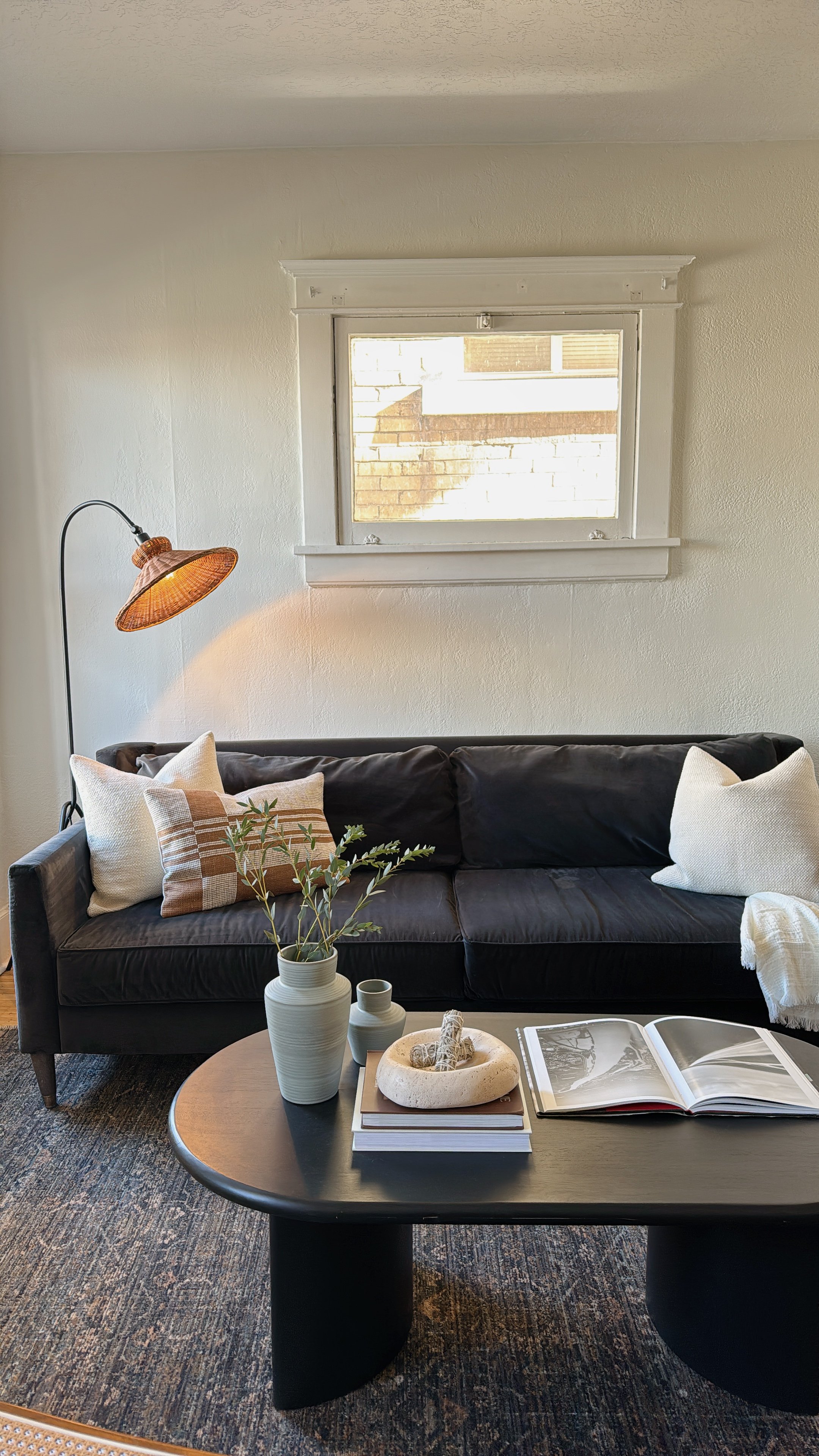 Living room with a black sofa, decorative pillows, a round black coffee table with vases and an open photo album, and a standing lamp with a woven shade near a window with white trim.