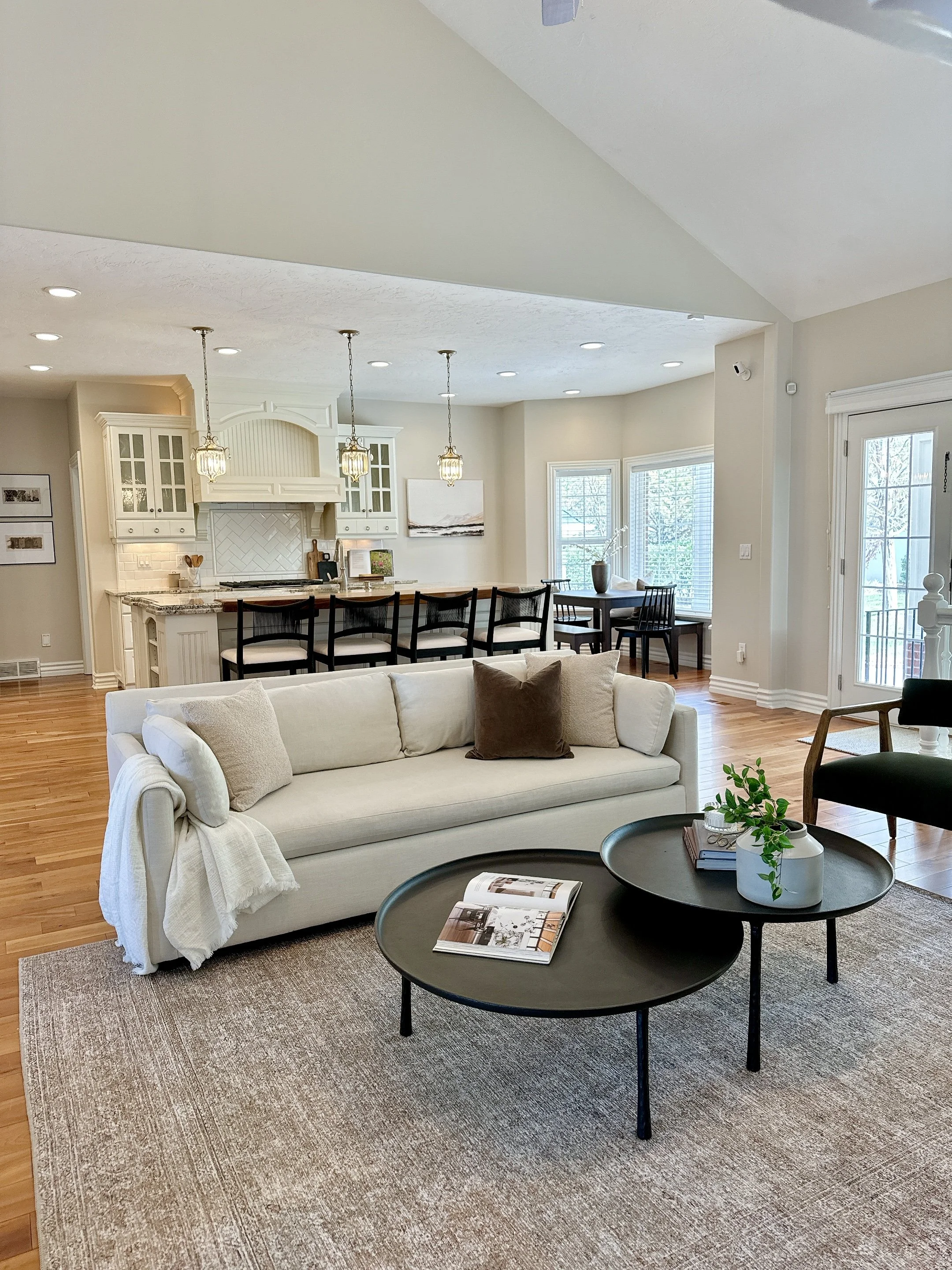 Staged living room with a white sofa, beige pillows, black coffee tables, a beige area rug, and a view of the kitchen and dining area with bar stools, pendant lights, and windows with natural light by Riley James Interiors.