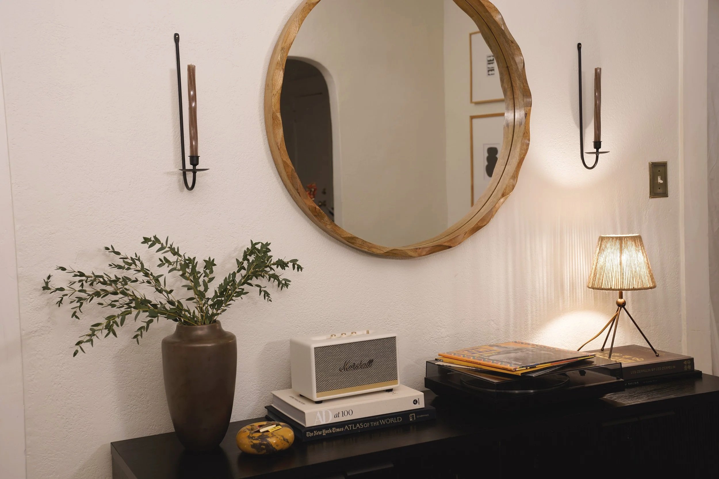 Living room wall with round wooden mirror, wall sconces, a dark sideboard with a vase of green foliage, a small Marshall speaker, stacked books, and vinyl records illuminated by a table lamp.
