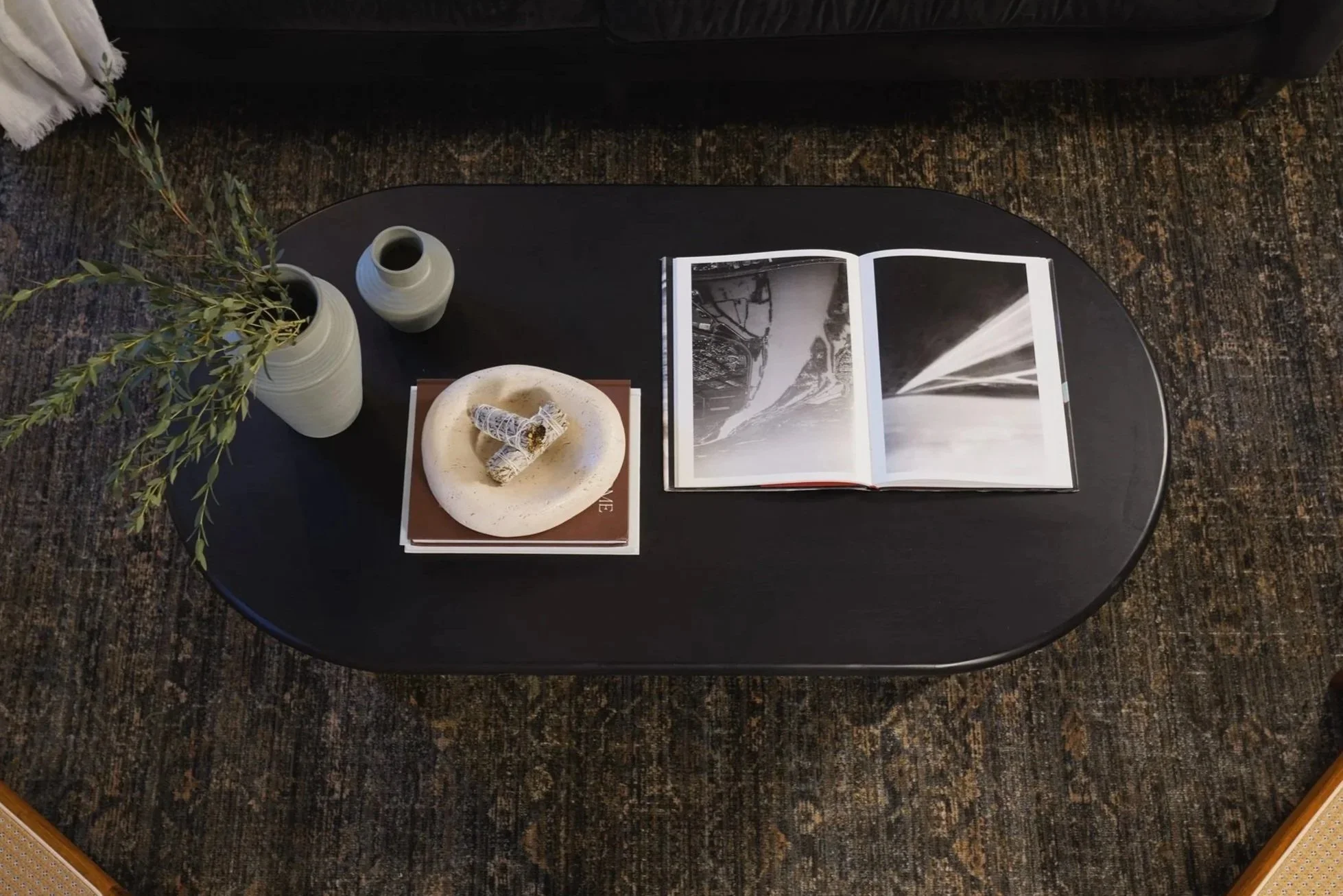 Black oval coffee table with vases, candles, books, an open photo album, and a small decorative object on top, placed on a dark patterned rug
