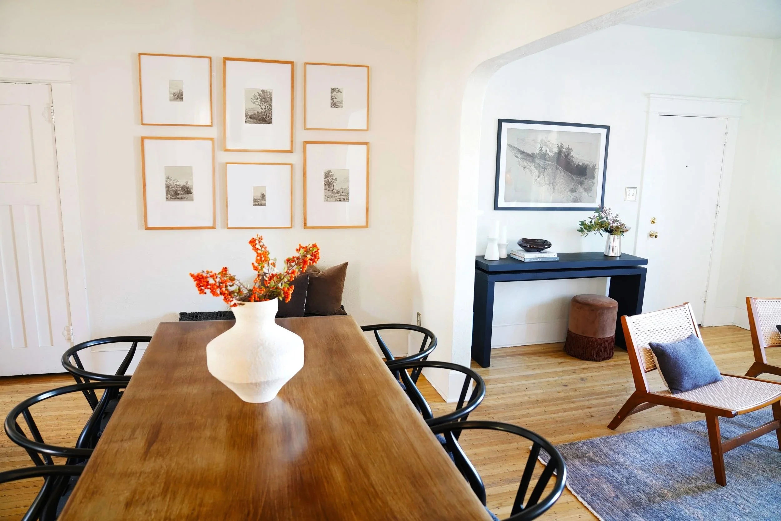A dining room with a wooden table, black chairs, a white vase with orange flowers, framed artwork on the walls, a small black table with decorative objects, a brown stool, and a chair with a pillow, all on a wooden floor.