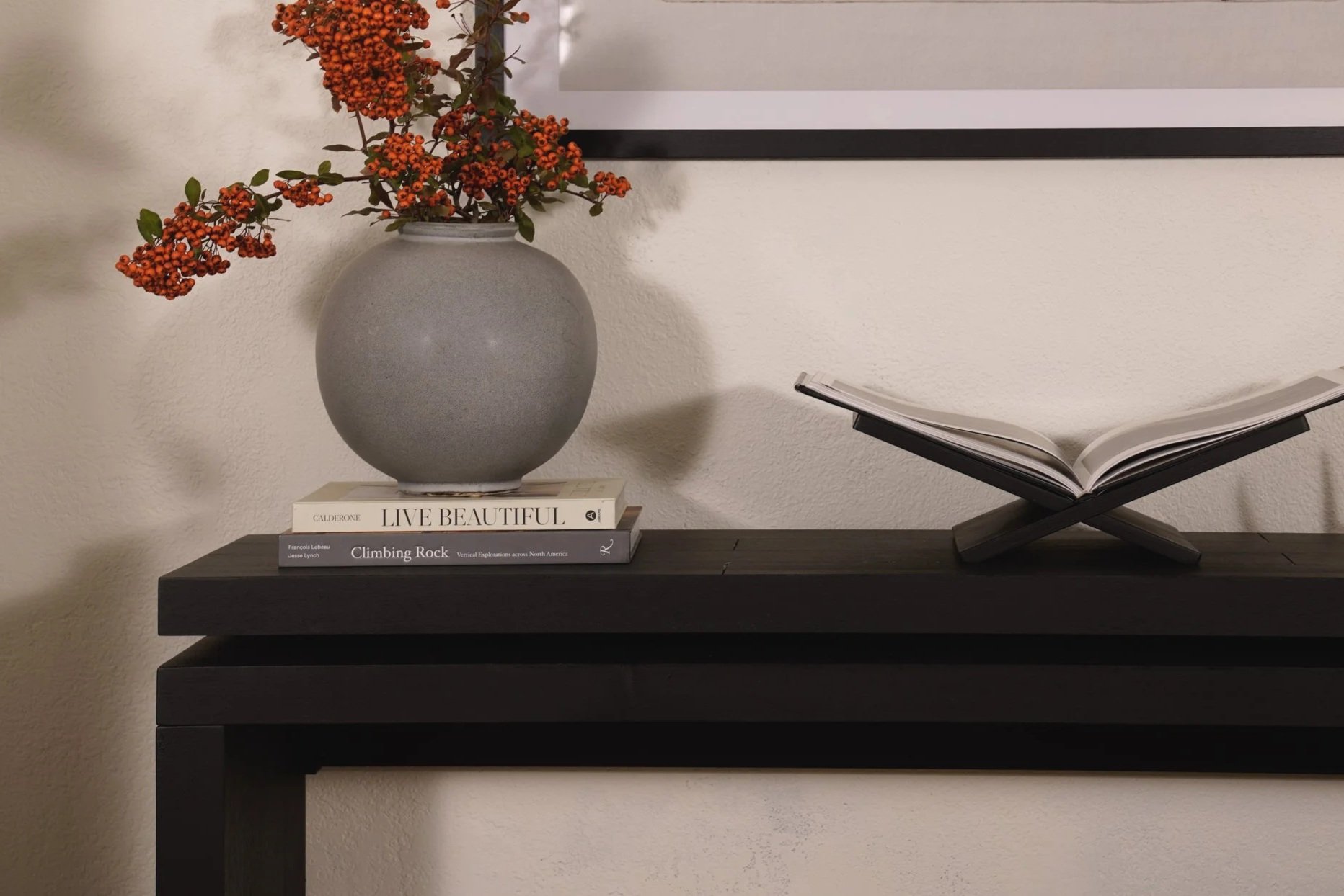A black table with a gray round vase containing orange berries and green leaves. Three books are stacked under the vase, and an open book is on a black stand to the right. A framed picture hangs on the white wall behind.