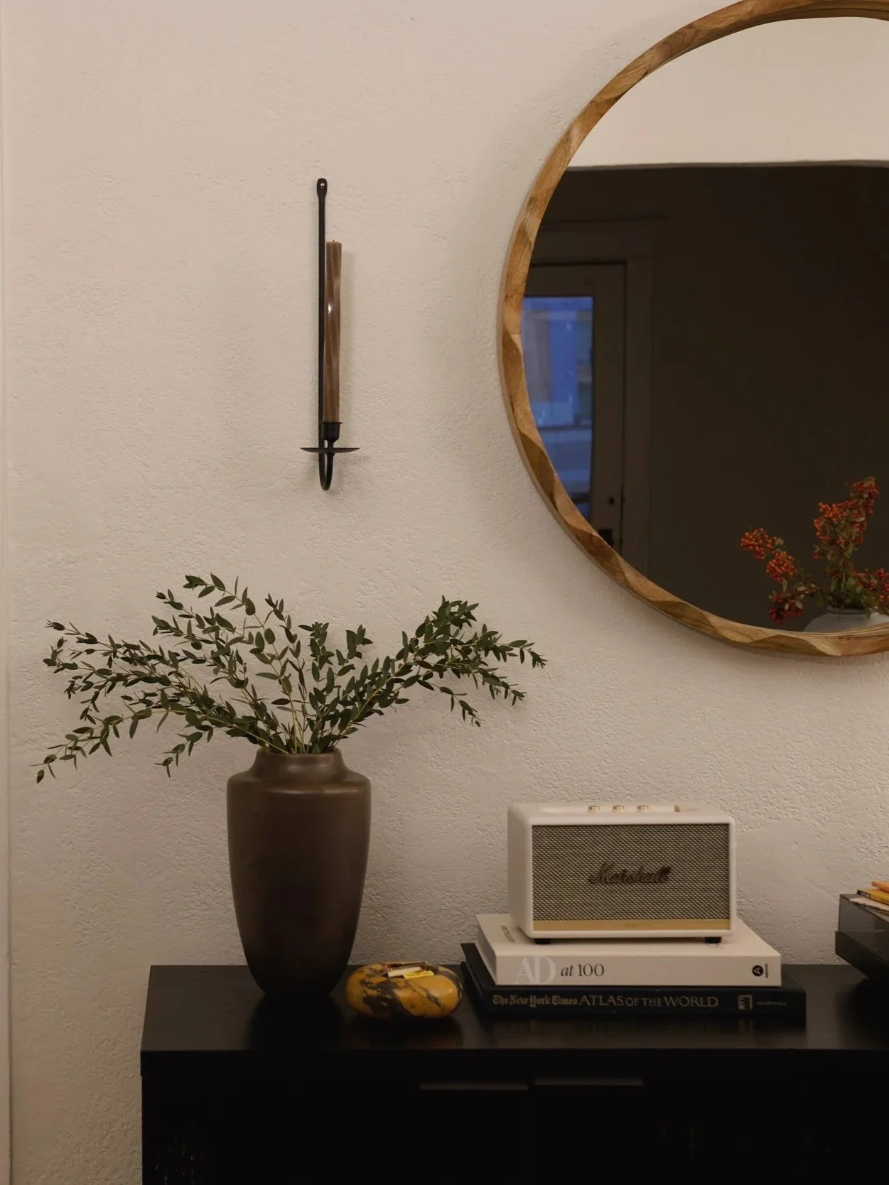 A black console table holding a tall brown vase with green leafy branches, a yellow decorative object, a stack of books, a small whitemarshall speaker, and a round mirror with a wooden frame hanging on a beige wall behind