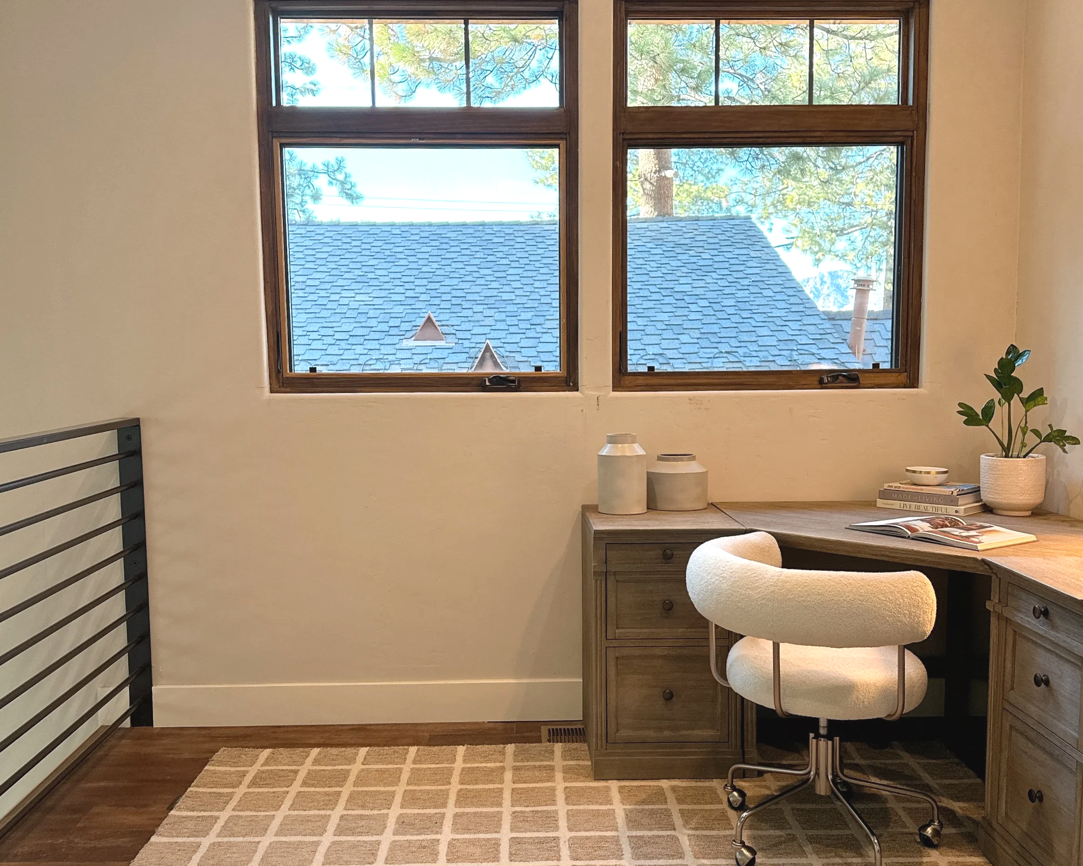 A cozy home office corner with a wooden desk, a plush white desk chair, decorative jars, a potted plant, and books. Behind the desk are two large windows showing a view of a roof, trees, and the sky.