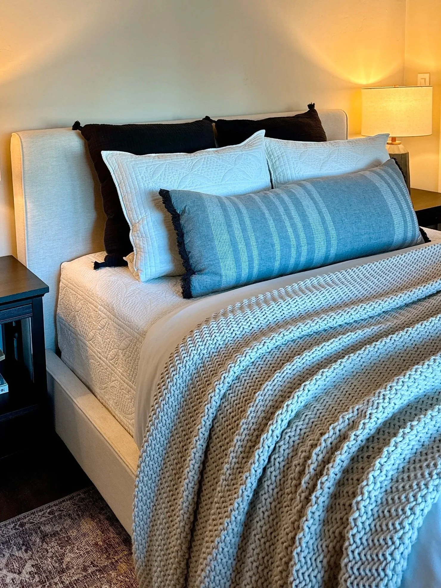 A neatly made bed with a beige upholstered headboard, layered with pillows in black, white, and blue with different textures, a cream quilt, and a chunky knit beige blanket, flanked by dark wood nightstands with a lit table lamp on one side.